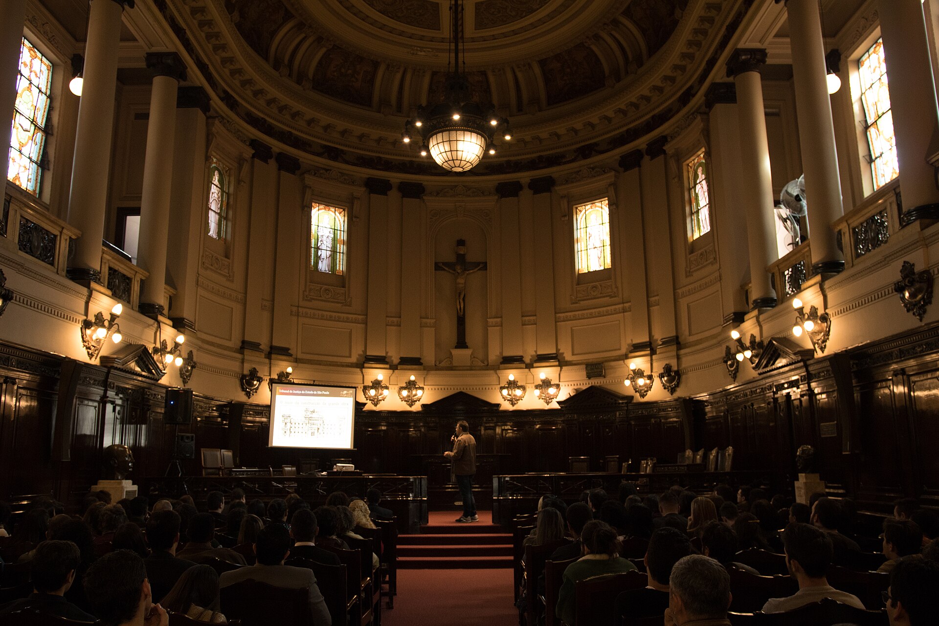 A domed hall with stained-glass windows, a crucifix and an audience seated before a speaker inside the Palacio da Justica in Sao Paulo