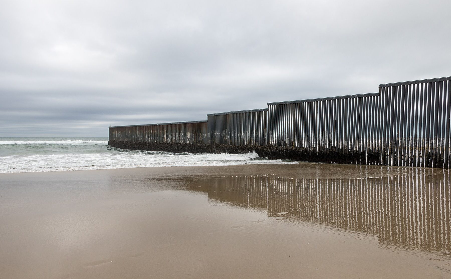 A tall steel-bar border wall runs down a grey beach into the sea under an overcast sky, reflected in wet sand