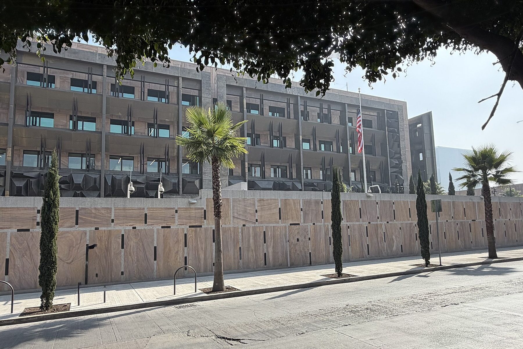 The new US Embassy compound in Mexico City seen from the street, with a US flag above a stone perimeter wall and palm trees