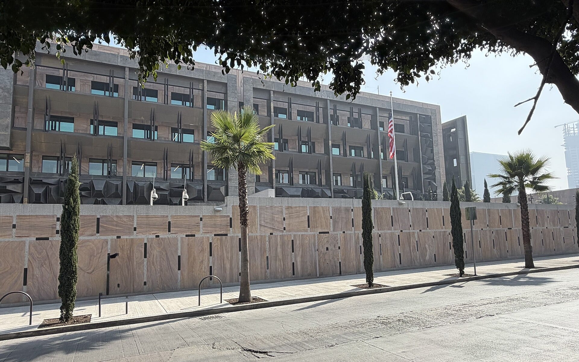The new US Embassy compound in Mexico City seen from the street, with a US flag above a stone perimeter wall and palm trees