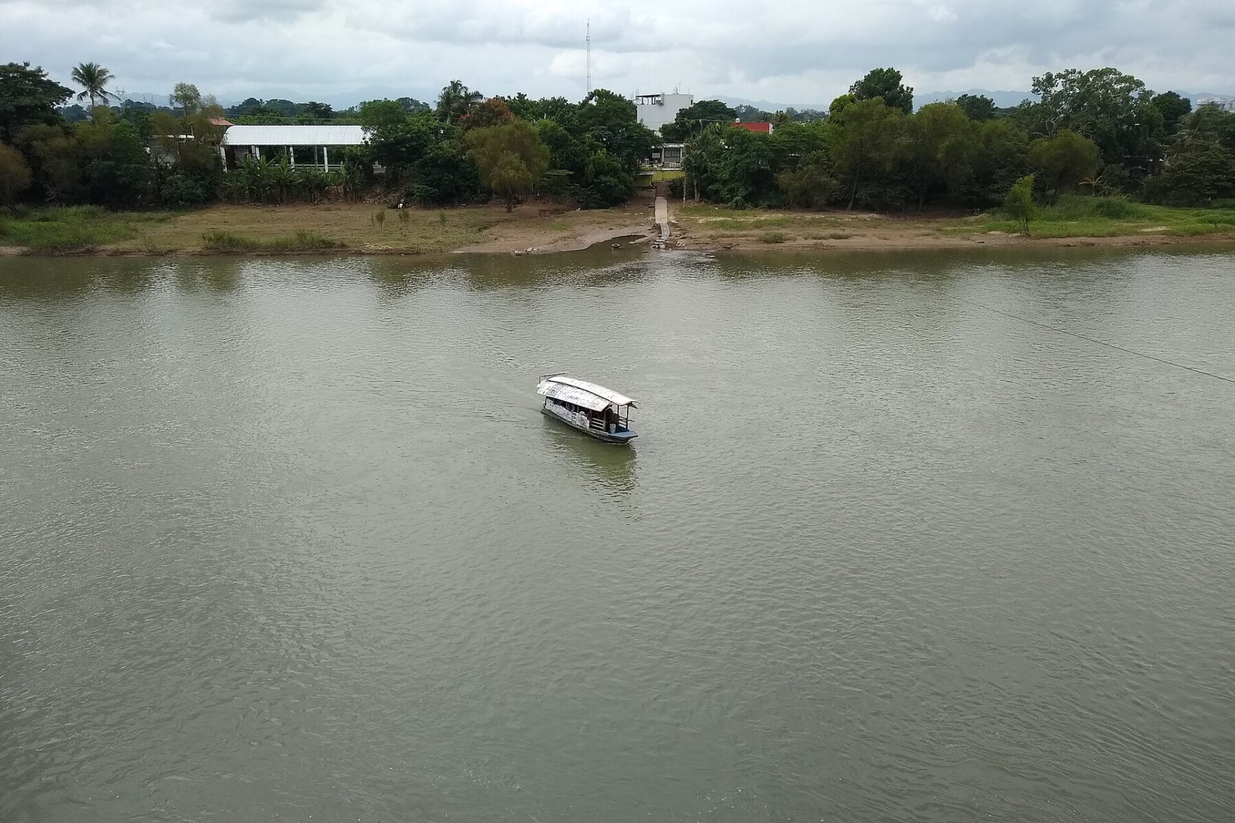 A small covered passenger boat crosses the wide, green Papaloapan River in Tuxtepec, with trees and low buildings on the far bank