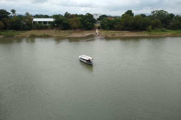 A small covered passenger boat crosses the wide, green Papaloapan River in Tuxtepec, with trees and low buildings on the far bank