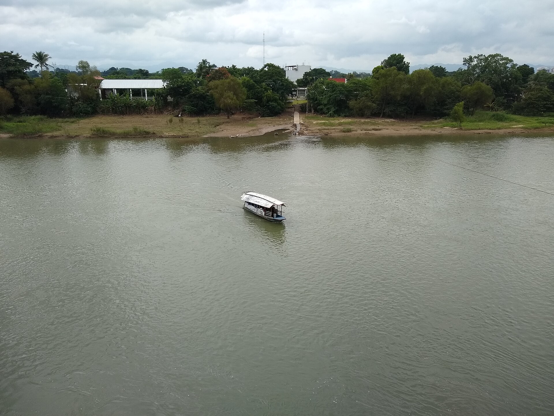 A small covered passenger boat crosses the wide, green Papaloapan River in Tuxtepec, with trees and low buildings on the far bank