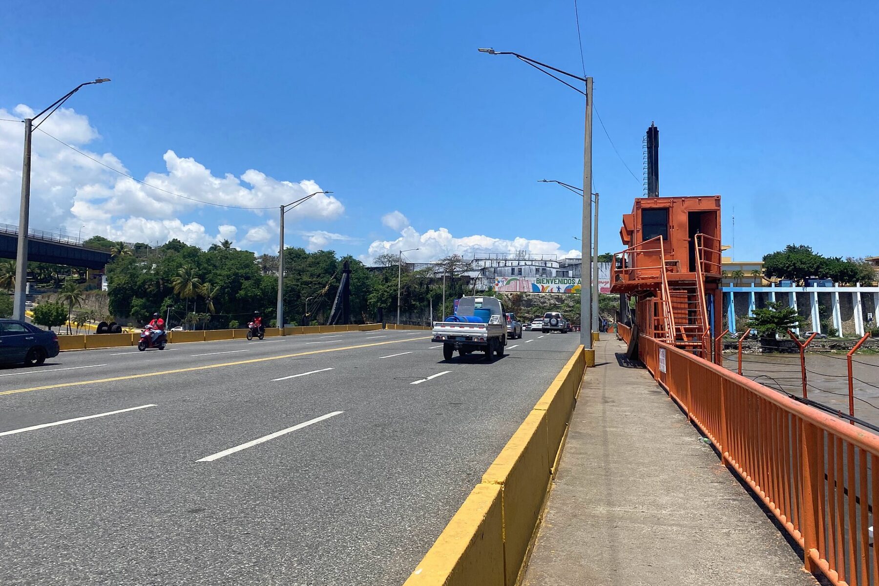 Traffic on a multi-lane road bridge under a blue sky, with an orange control cabin and railing on the right and street lights and trees ahead