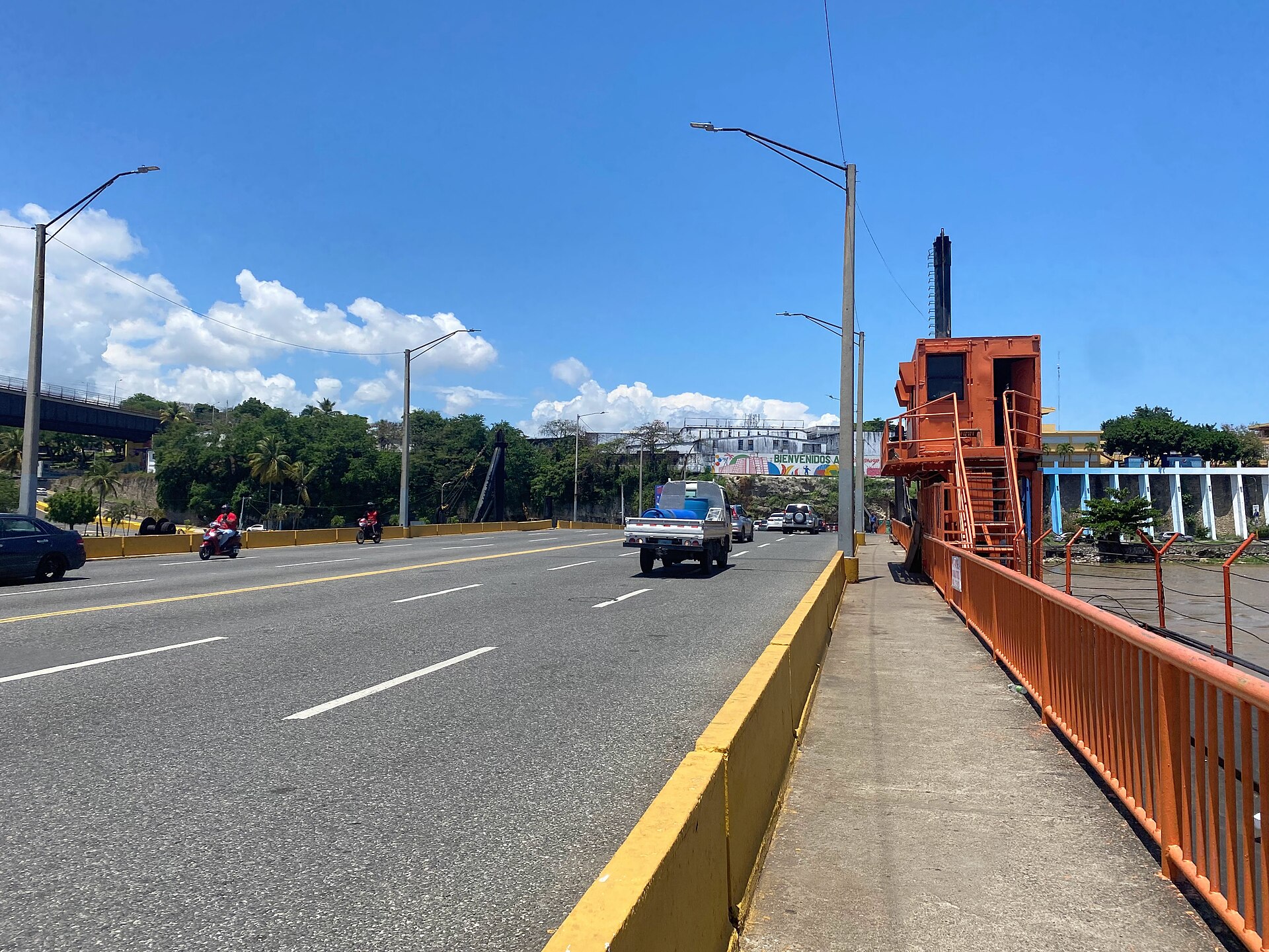 Traffic on a multi-lane road bridge under a blue sky, with an orange control cabin and railing on the right and street lights and trees ahead