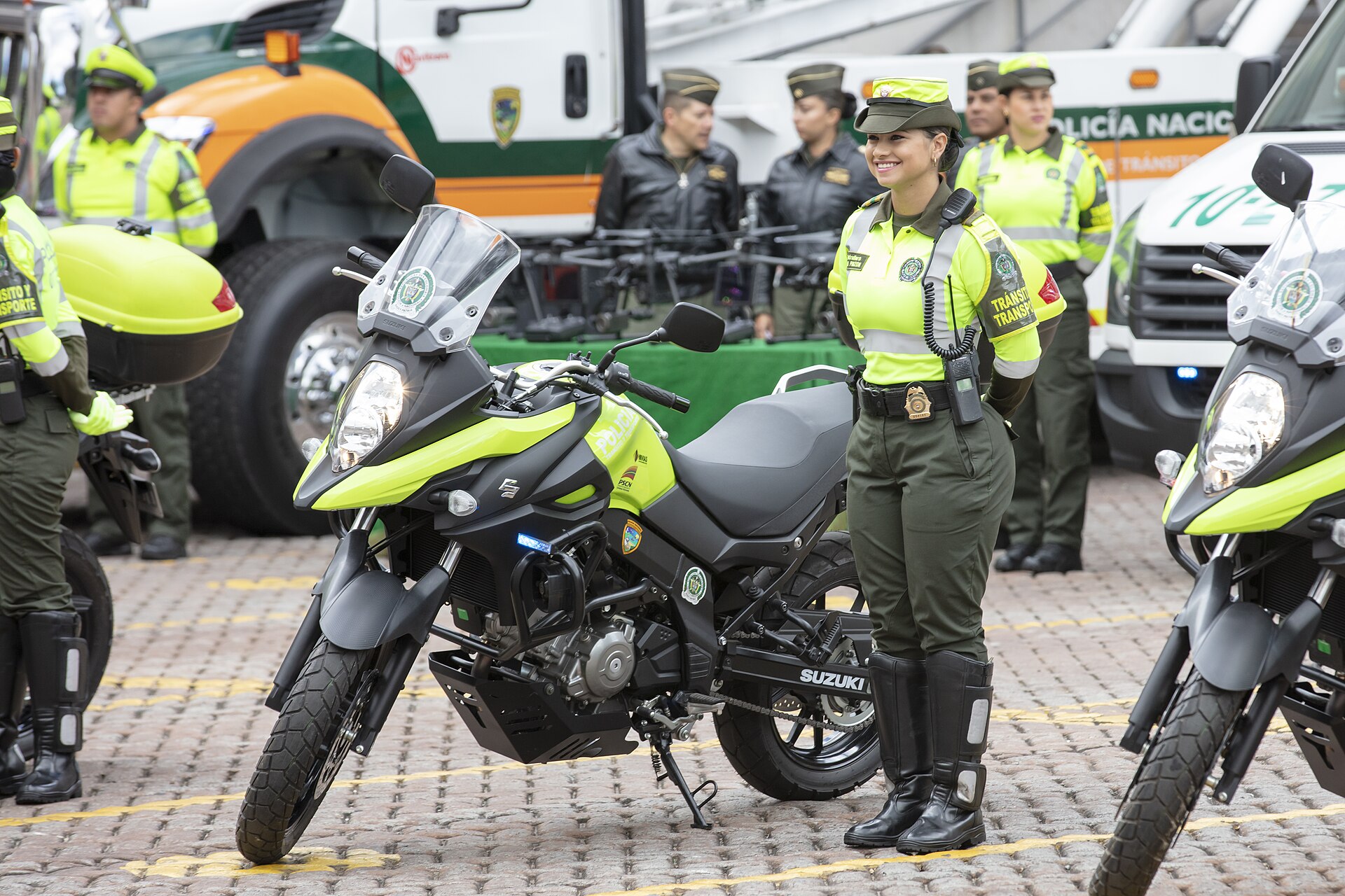 A Colombian National Police officer in a green and yellow vest stands beside a police motorcycle, with other officers and police vehicles behind
