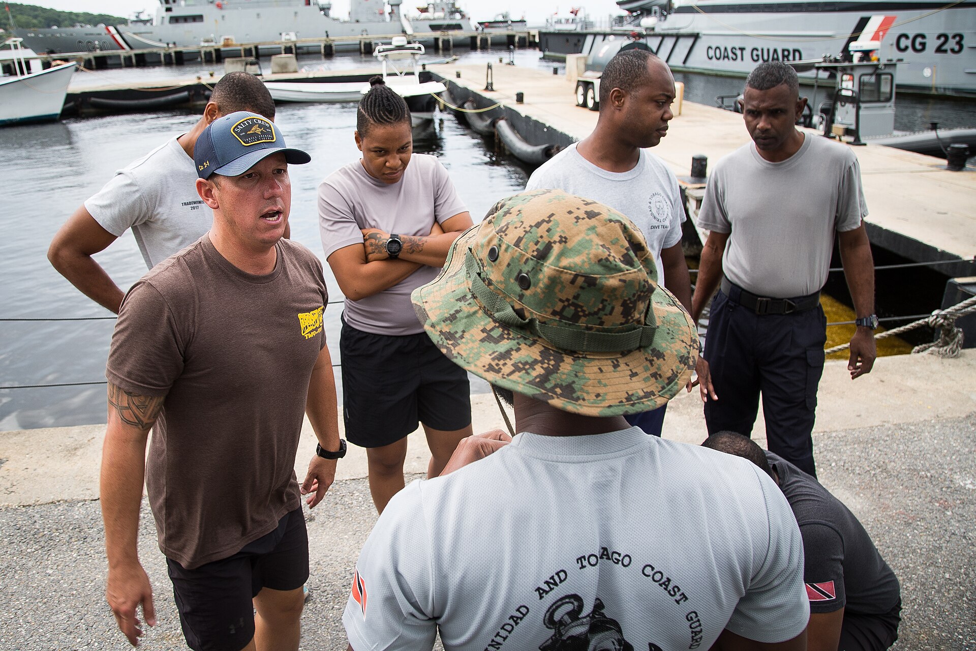 A US Navy diver in a cap speaks to Trinidad and Tobago Coast Guard personnel on a quay beside Coast Guard vessels
