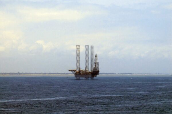 A jack-up offshore oil drilling rig standing in calm dark blue water under a pale cloudy sky, with a low coastline behind it