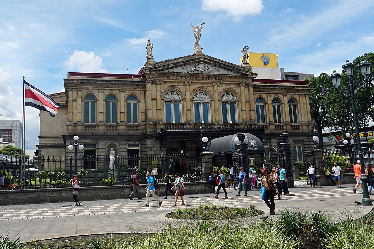 Main facade of the Teatro Nacional in central San José, Costa Rica, with the national flag at left and pedestrians crossing the plaza.