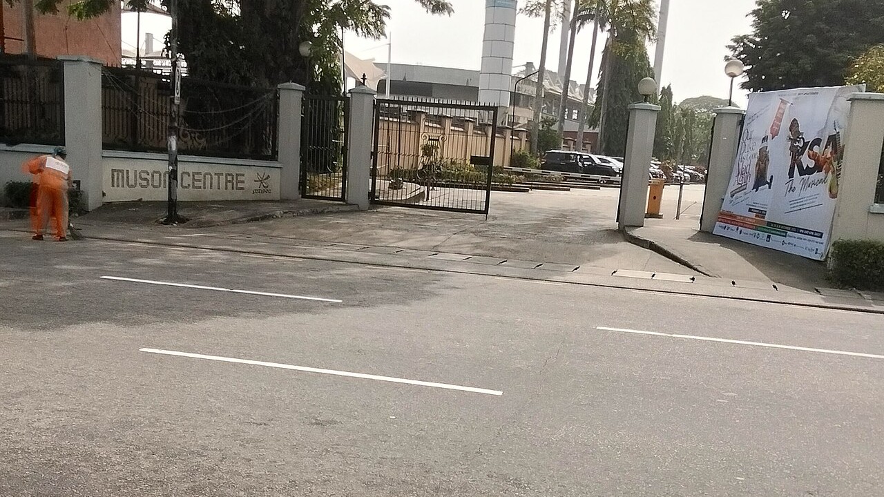 Entrance gate of the MUSON Centre on a Lagos street, with its name on the wall at left and a show banner at right.