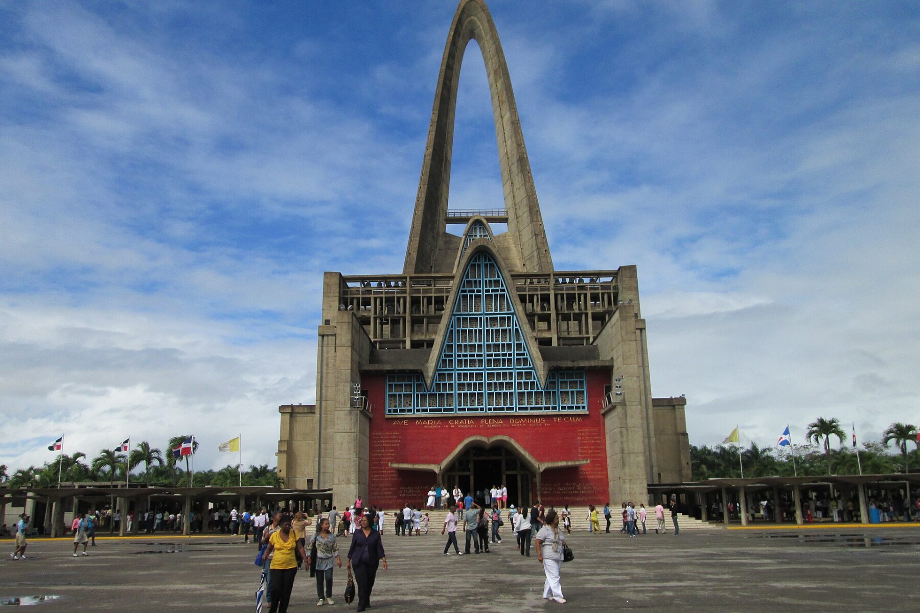 Basilica Cathedral of Our Lady of Altagracia in Higüey, a tall concrete arch above a red facade, with visitors crossing the forecourt