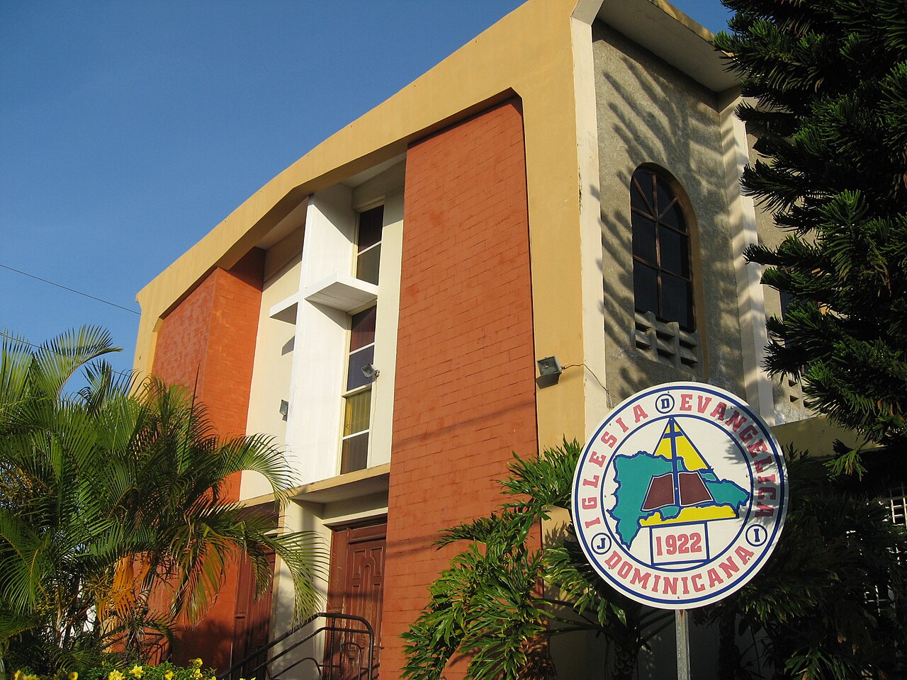 Brick and plaster church building with a sign reading Iglesia Evangélica Dominicana 1922 beside palms