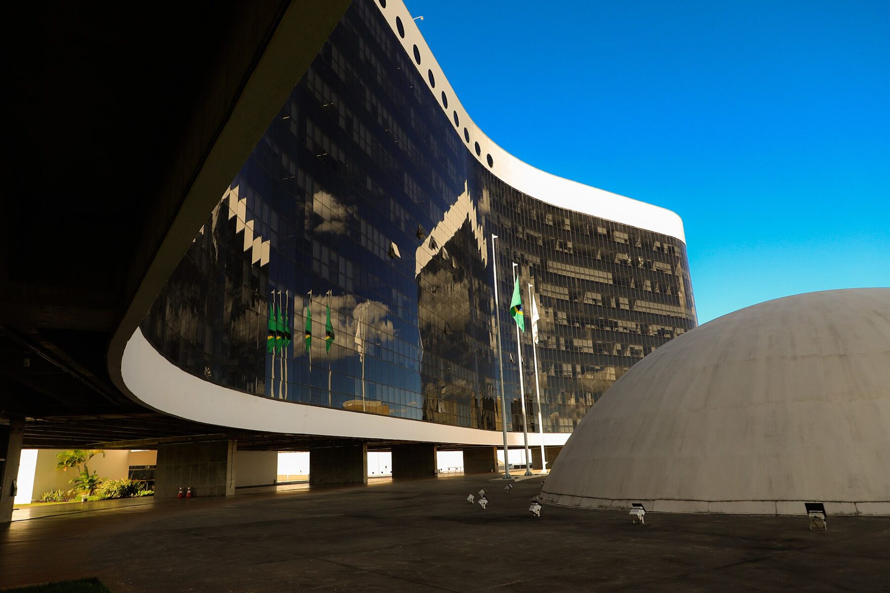 The curved glass facade of the Superior Electoral Court building in Brasília reflects Brazilian flags, beside a white concrete dome under a blue sky