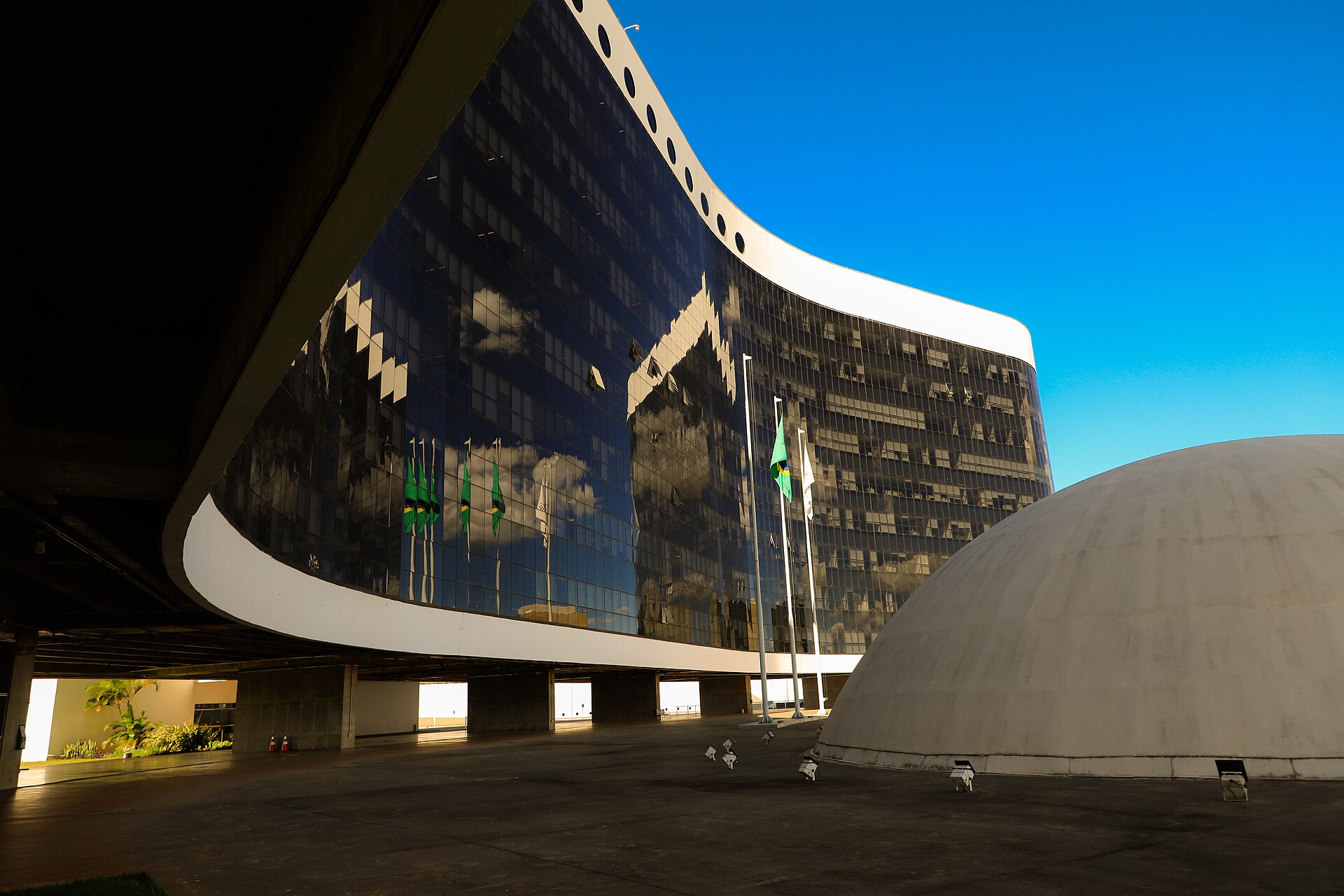 The curved glass facade of the Superior Electoral Court building in Brasília reflects Brazilian flags, beside a white concrete dome under a blue sky