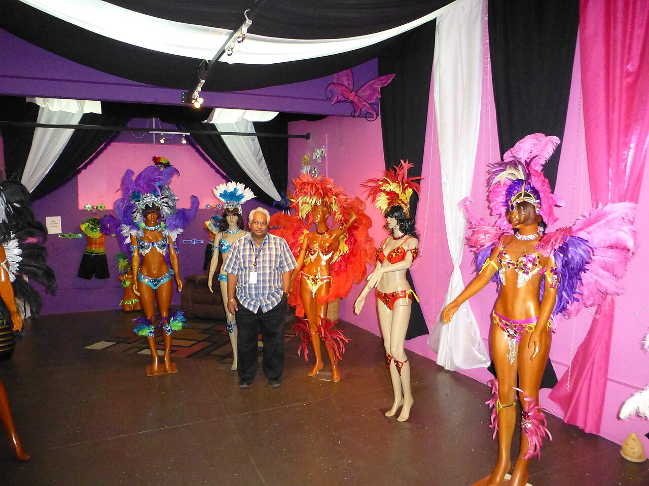 Mannequins in feathered, sequinned Carnival costumes in pink, purple, orange and red on display in a draped room, with a man in a checked shirt among them