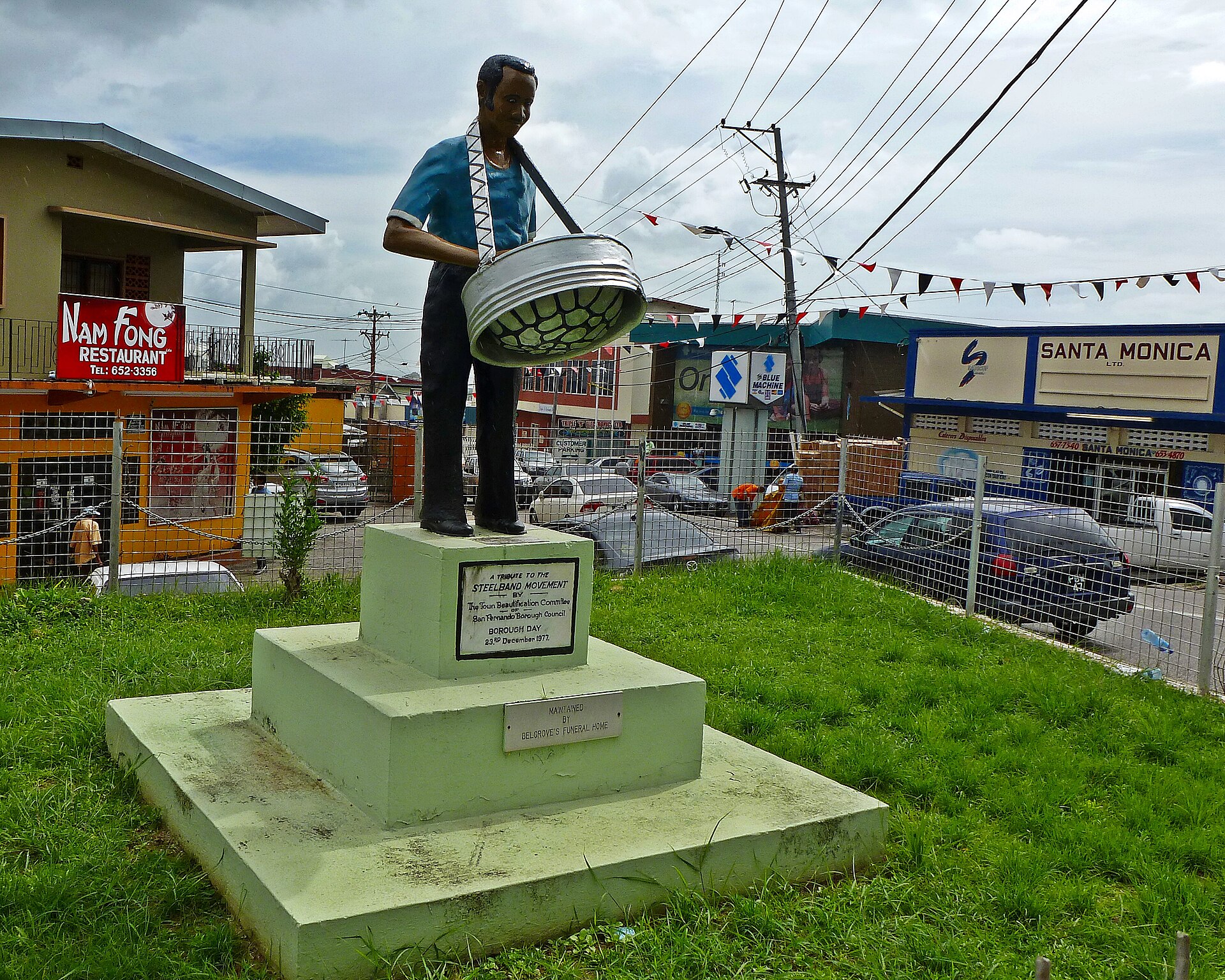 A painted statue of a man playing a steel pan stands on a pale green plinth with a plaque, beside a street with shops and parked cars