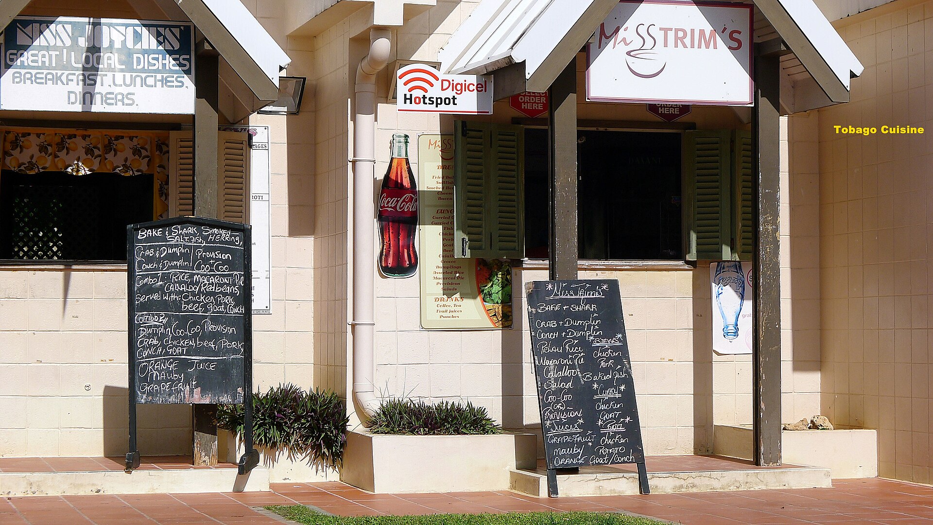Two small cook shops with chalkboard menus listing crab and dumpling, bake and shark and other local dishes, under sloping roofs in Tobago