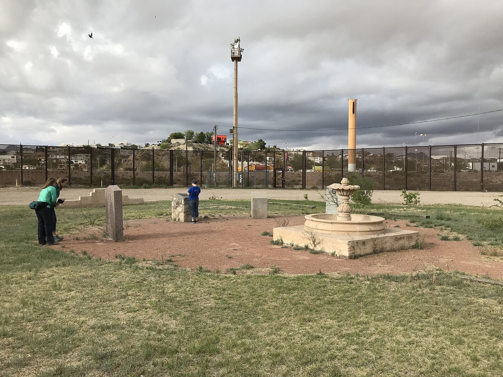 A stone fountain and markers at the Onate Crossing in El Paso with visitors, and the border fence with Ciudad Juarez hills behind