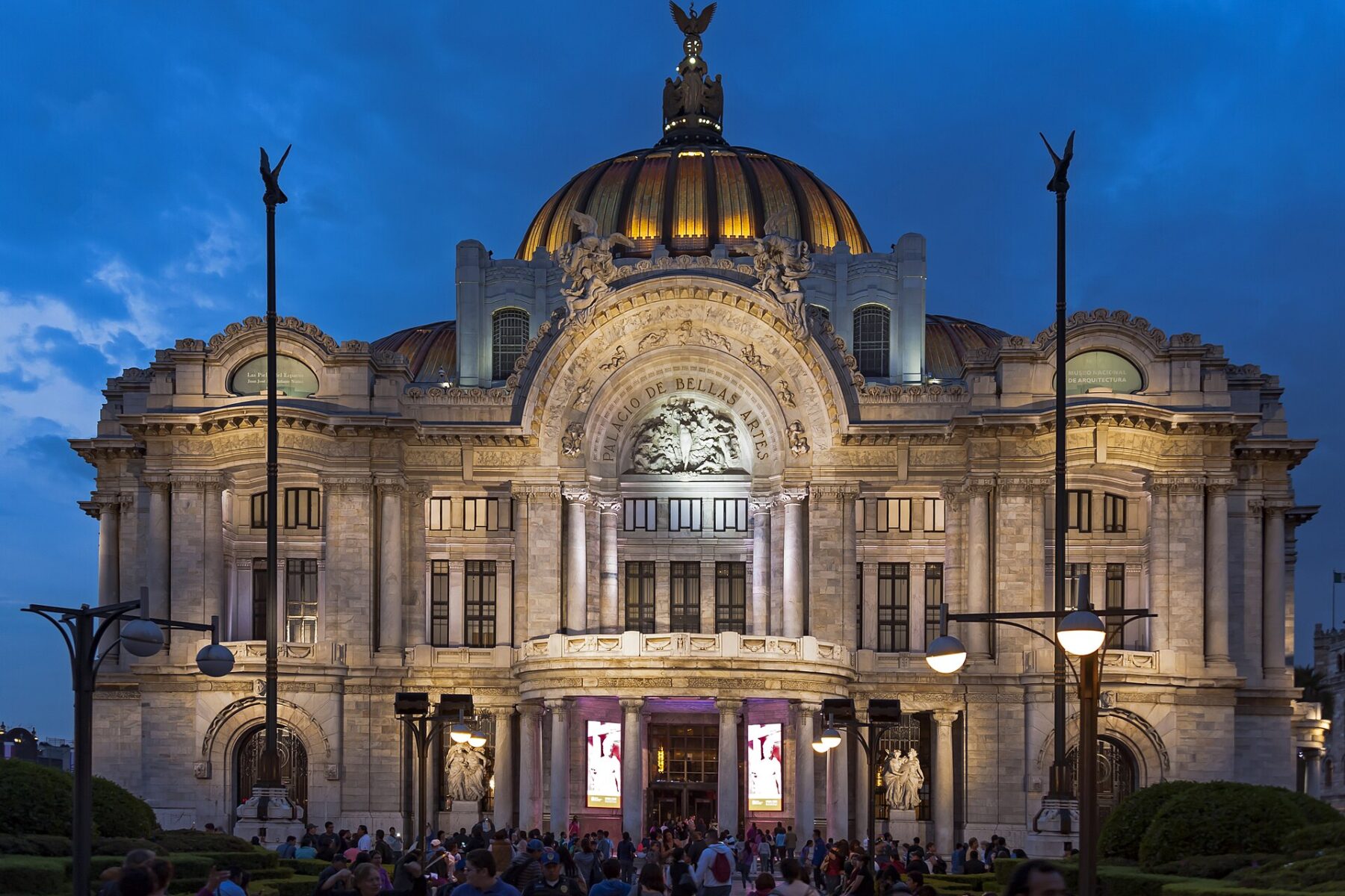 The marble facade and golden dome of the Palacio de Bellas Artes in Mexico City lit up at dusk, with crowds on the plaza in front
