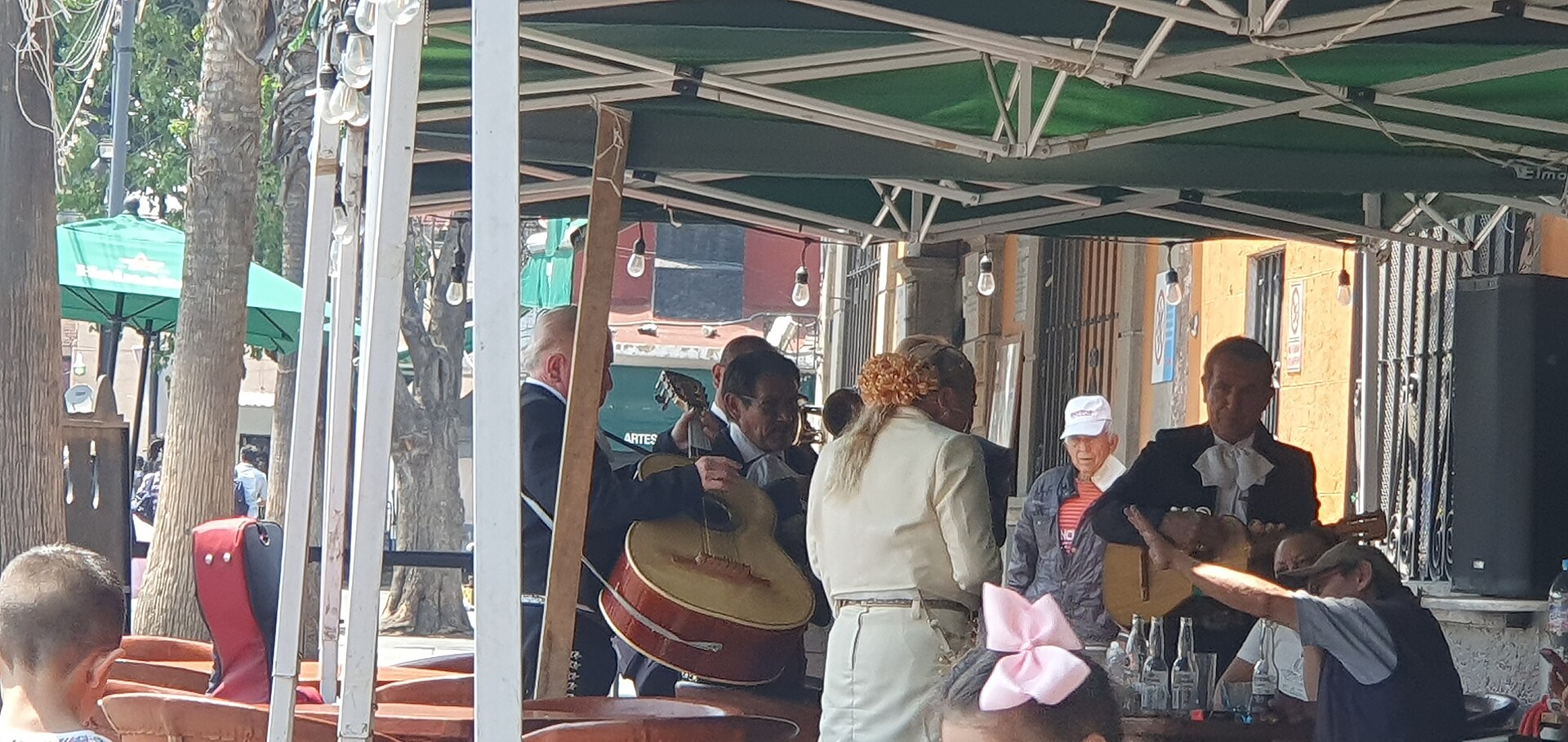 Mariachi musicians, most in dark charro suits and one woman in cream, play guitars and a large guitarrón for customers under a green awning at Plaza Garibaldi