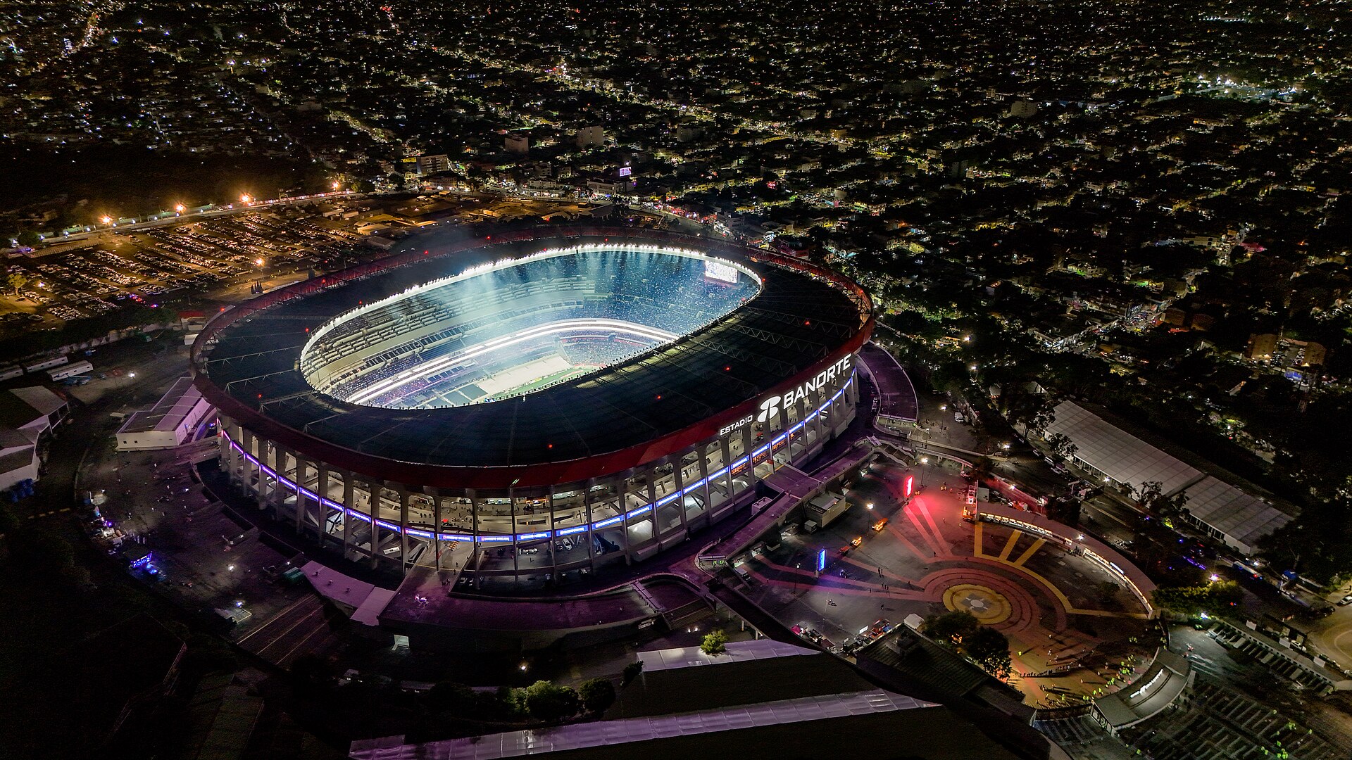 Night aerial view of the floodlit Estadio Azteca, with Banorte lettering on its roof, surrounded by the lights of Mexico City