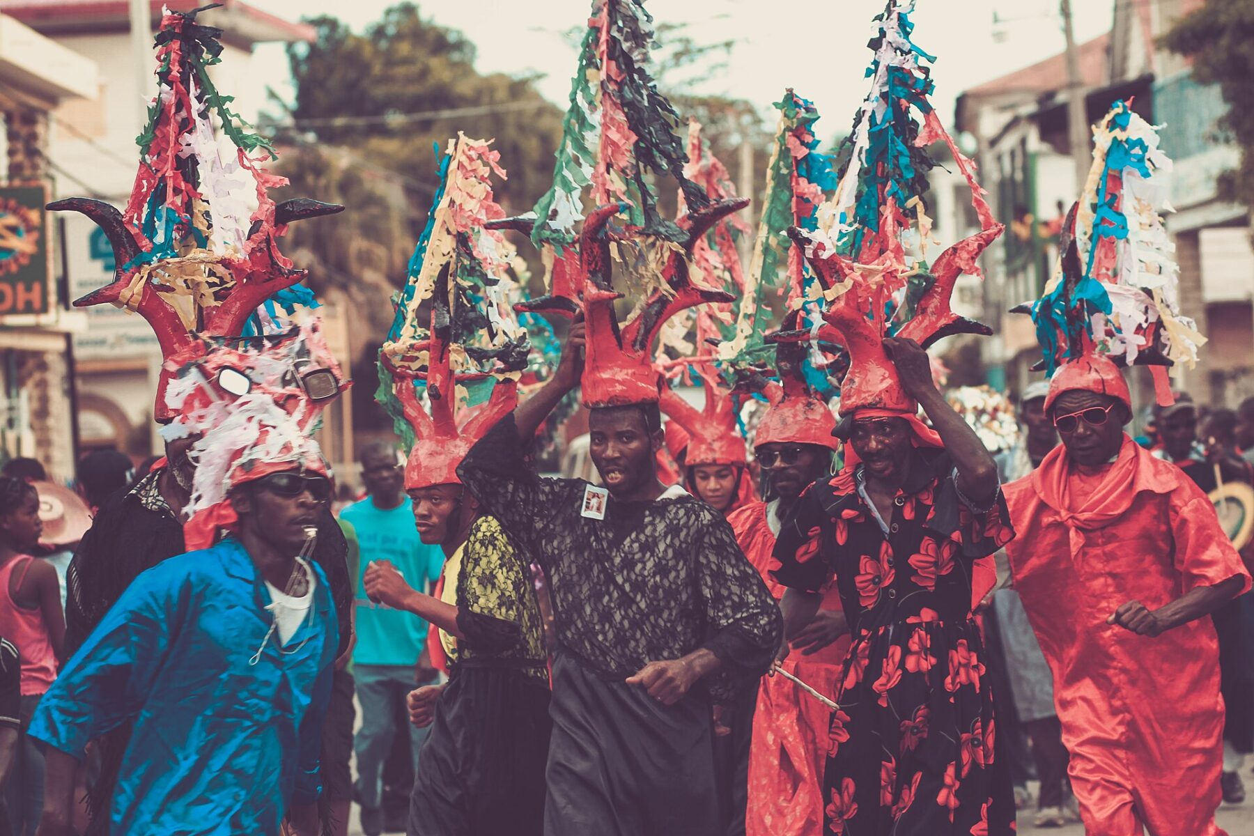 Marchers in tall red horned headdresses decorated with paper streamers parade through a street during the Jacmel carnival, with onlookers behind
