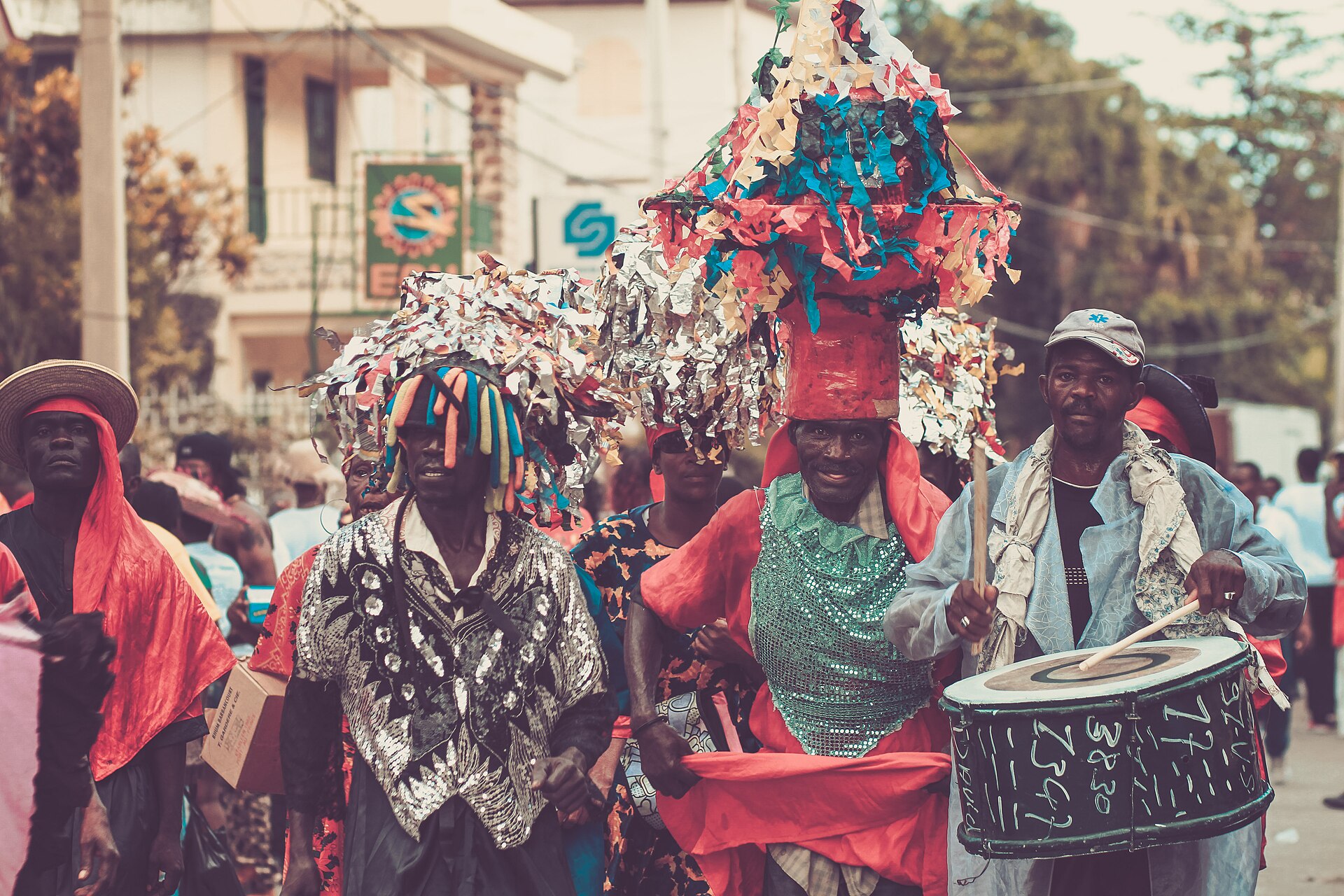 Costumed marchers in sequined shirts and paper headdresses walk a street while a man beats a painted drum