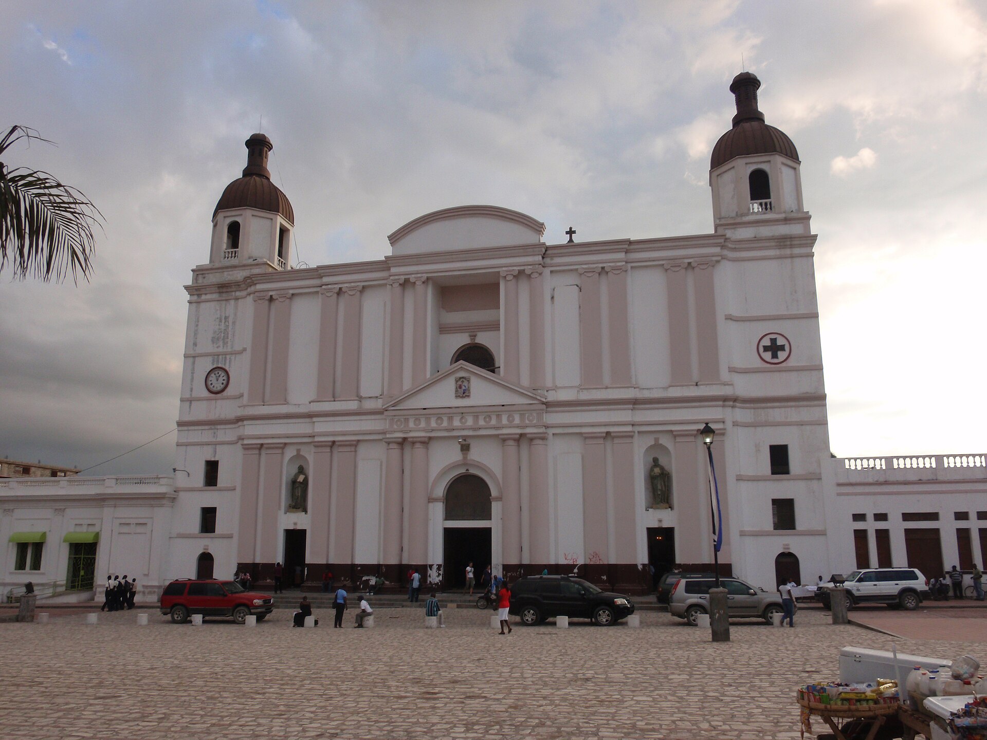 A pale two-towered cathedral facade facing a cobbled square with parked cars and people