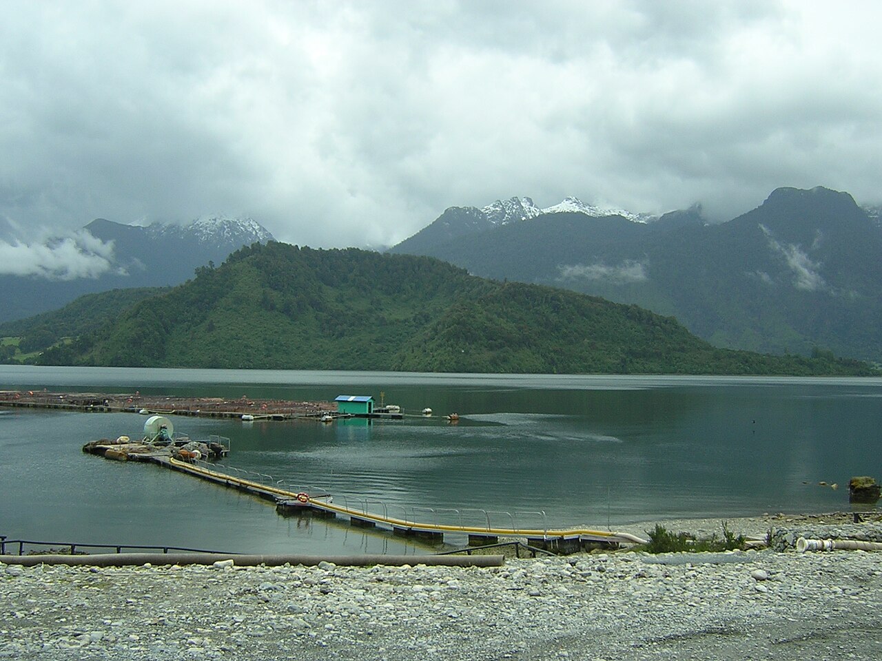 Floating walkway and pens of a salmon farm on a calm estuary beneath forested hills and cloud-covered mountains