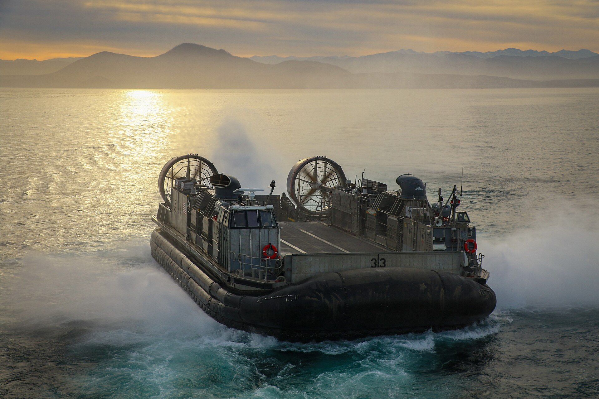 A US Navy hovercraft landing craft, number 33, speeding across water at sunset with mountains behind