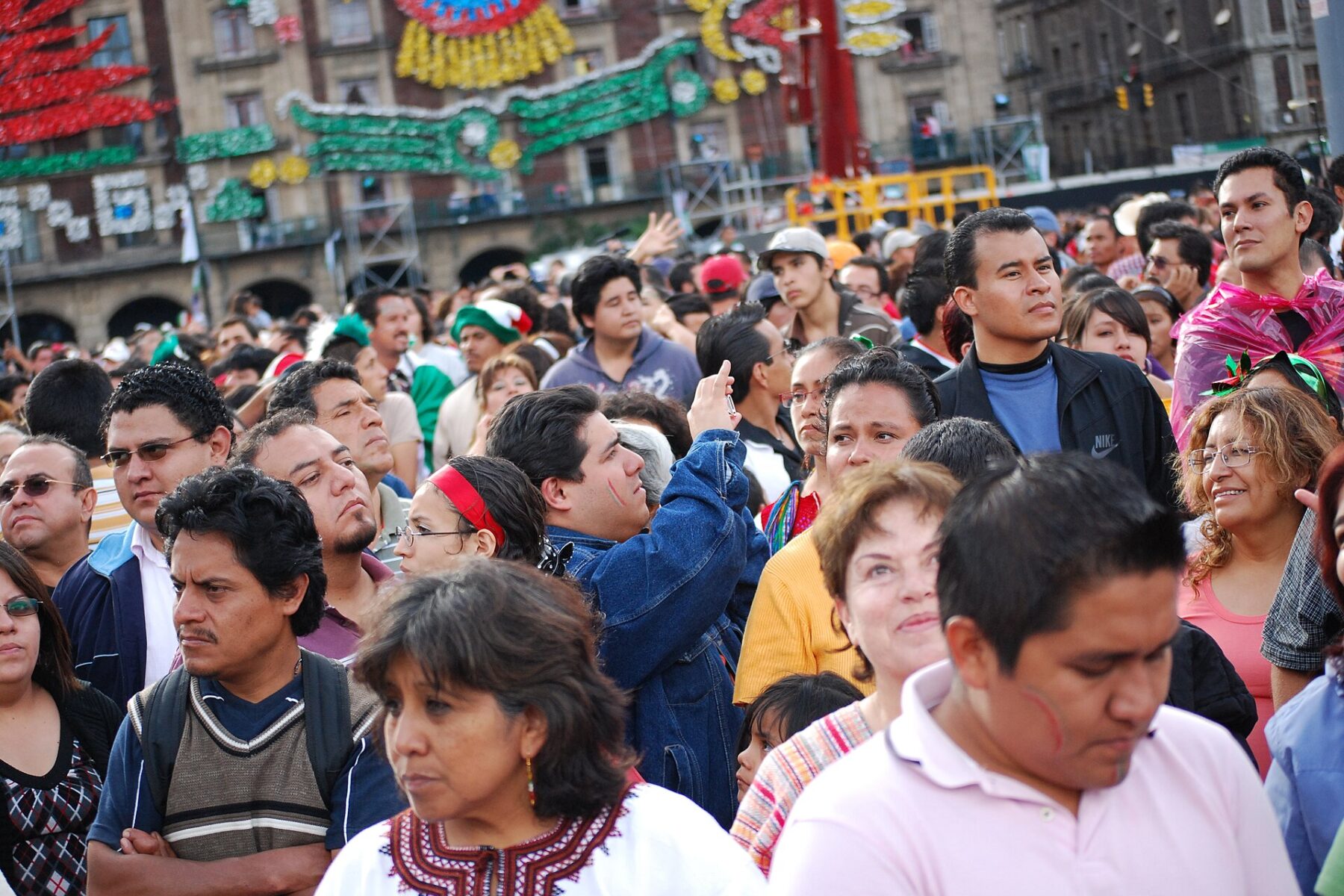 A dense crowd of Mexican men and women of different ages standing in Mexico City's Zócalo square, with patriotic decorations on the buildings behind