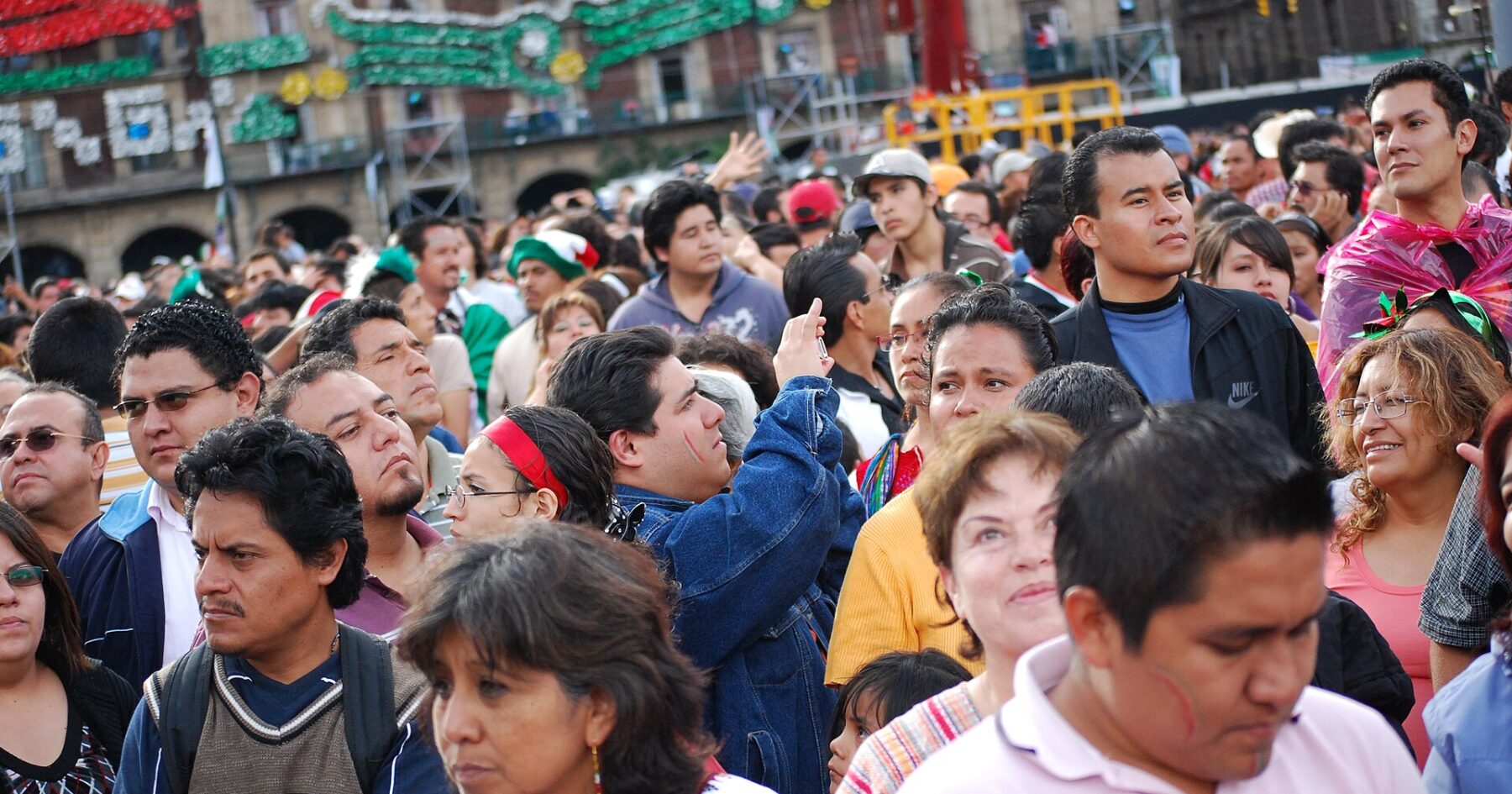 A dense crowd of Mexican men and women of different ages standing in Mexico City's Zócalo square, with patriotic decorations on the buildings behind