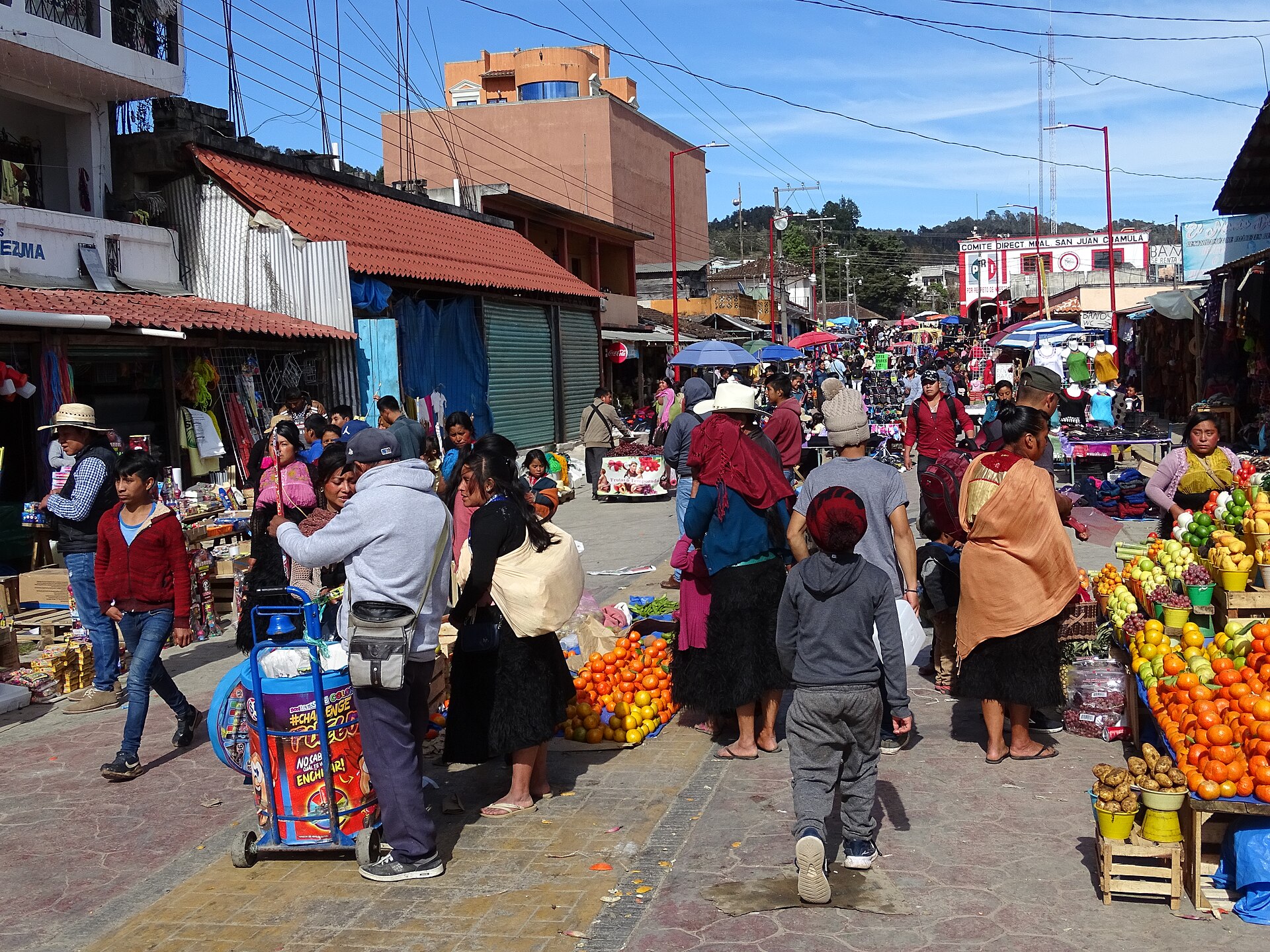 A busy market street in San Juan Chamula with fruit stalls, women in black skirts and coloured shawls, and shoppers walking past shops