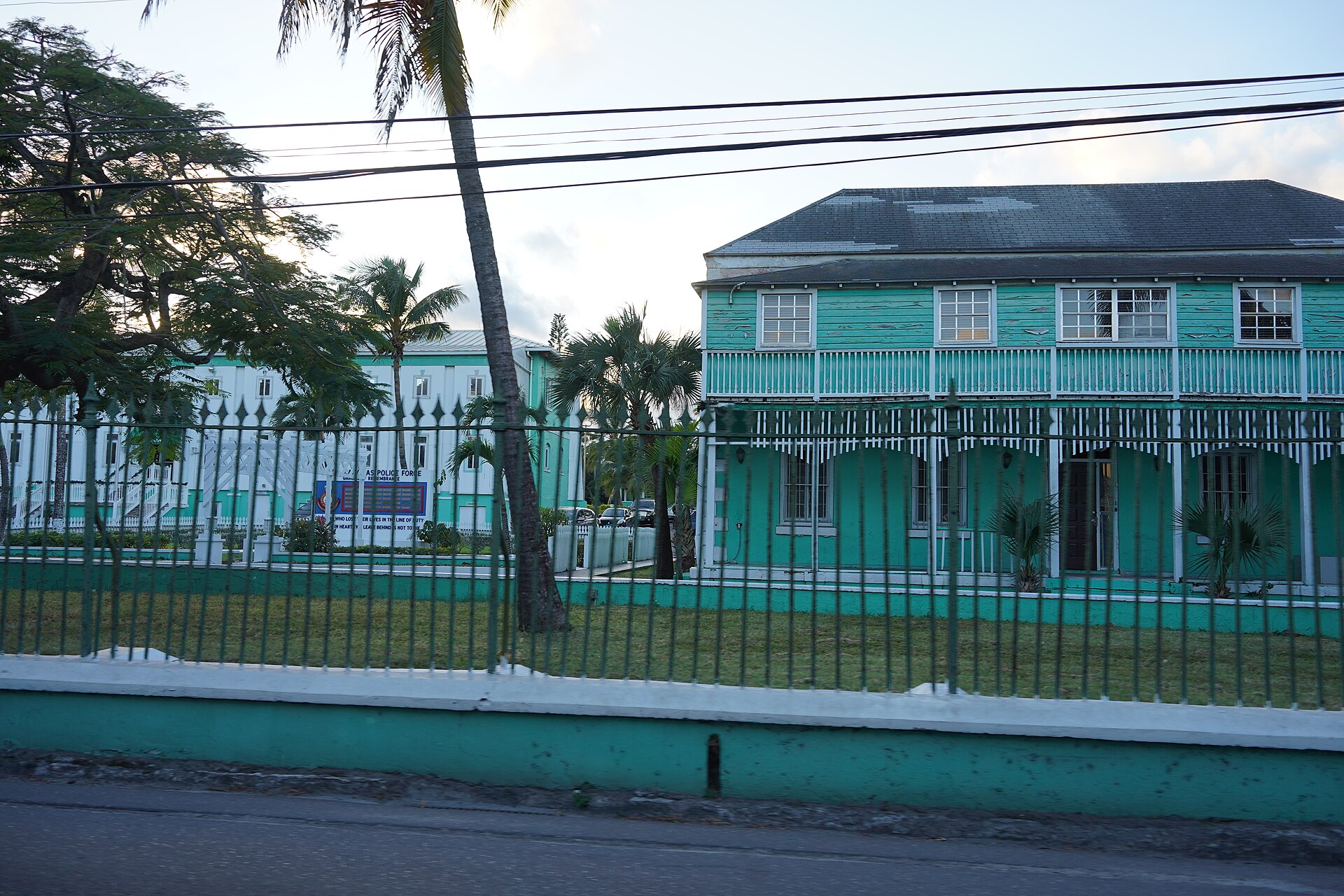 Teal-painted two-storey police headquarters building behind a green iron fence and palm trees in downtown Nassau