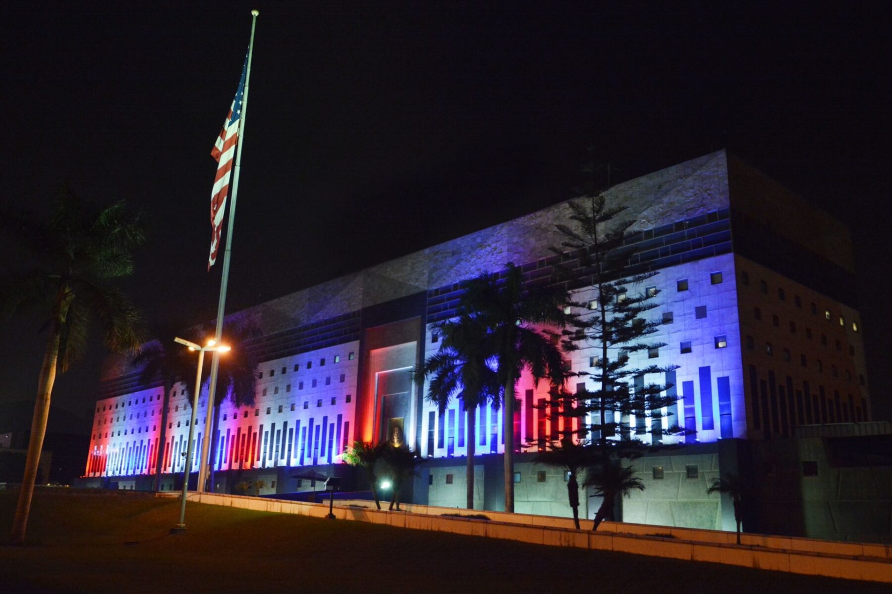 The large US Embassy building in Lima at night, its facade lit in red, white and blue, with a US flag on a pole and palm trees in front