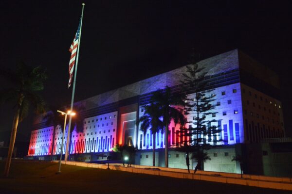 The large US Embassy building in Lima at night, its facade lit in red, white and blue, with a US flag on a pole and palm trees in front