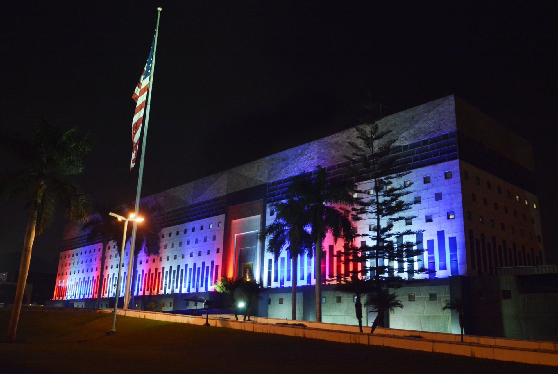 The large US Embassy building in Lima at night, its facade lit in red, white and blue, with a US flag on a pole and palm trees in front