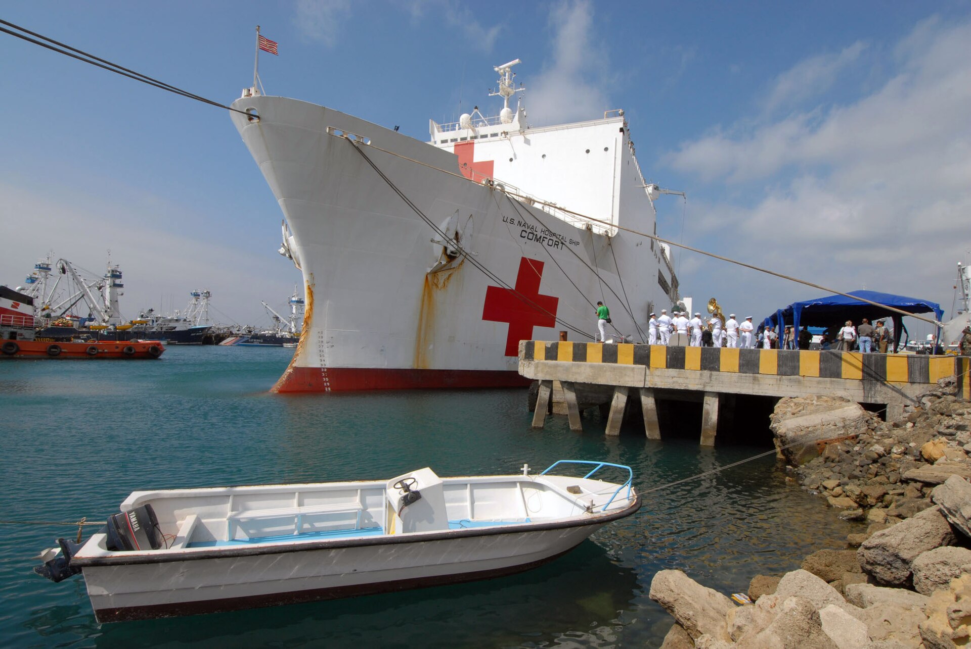 The white hospital ship USNS Comfort with a large red cross on its hull moored at a pier in Manta, Ecuador, with sailors on the pier and a small boat in the foreground