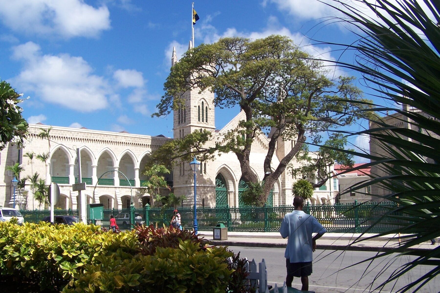 The Parliament Buildings in Bridgetown with the clock tower, a Barbados flag, a tree and a green iron fence under a blue sky