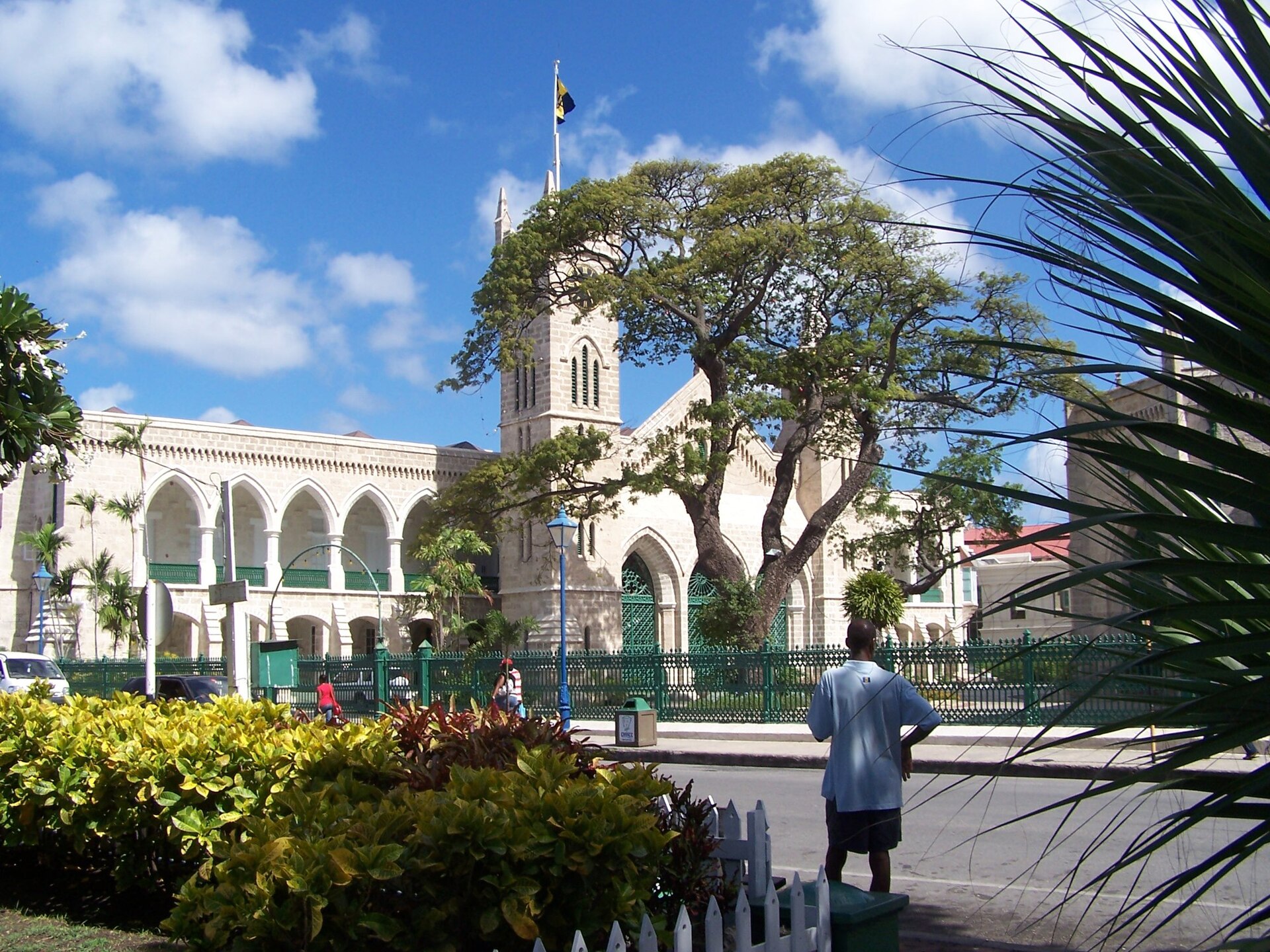 The Parliament Buildings in Bridgetown with the clock tower, a Barbados flag, a tree and a green iron fence under a blue sky