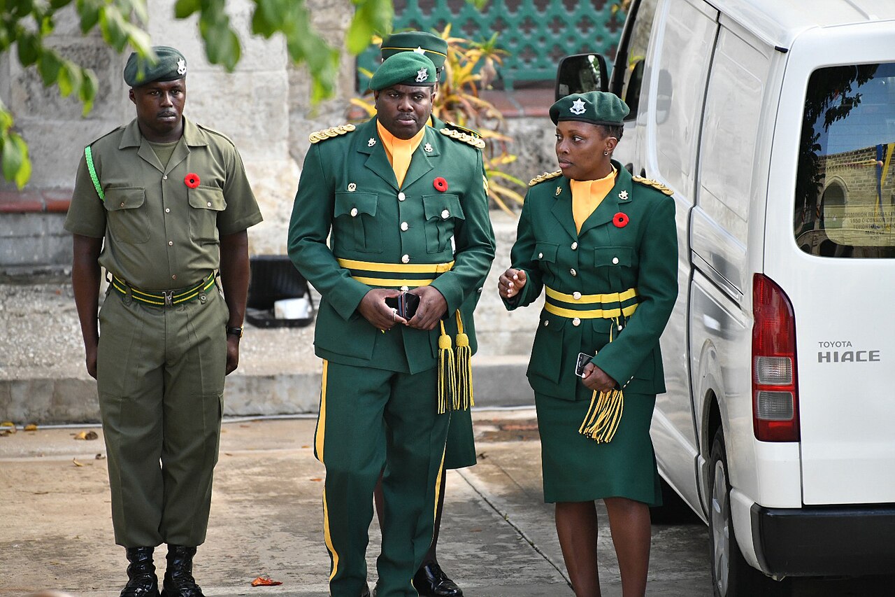 Three Barbados Defence Force members in green uniforms with red remembrance poppies standing beside a white van