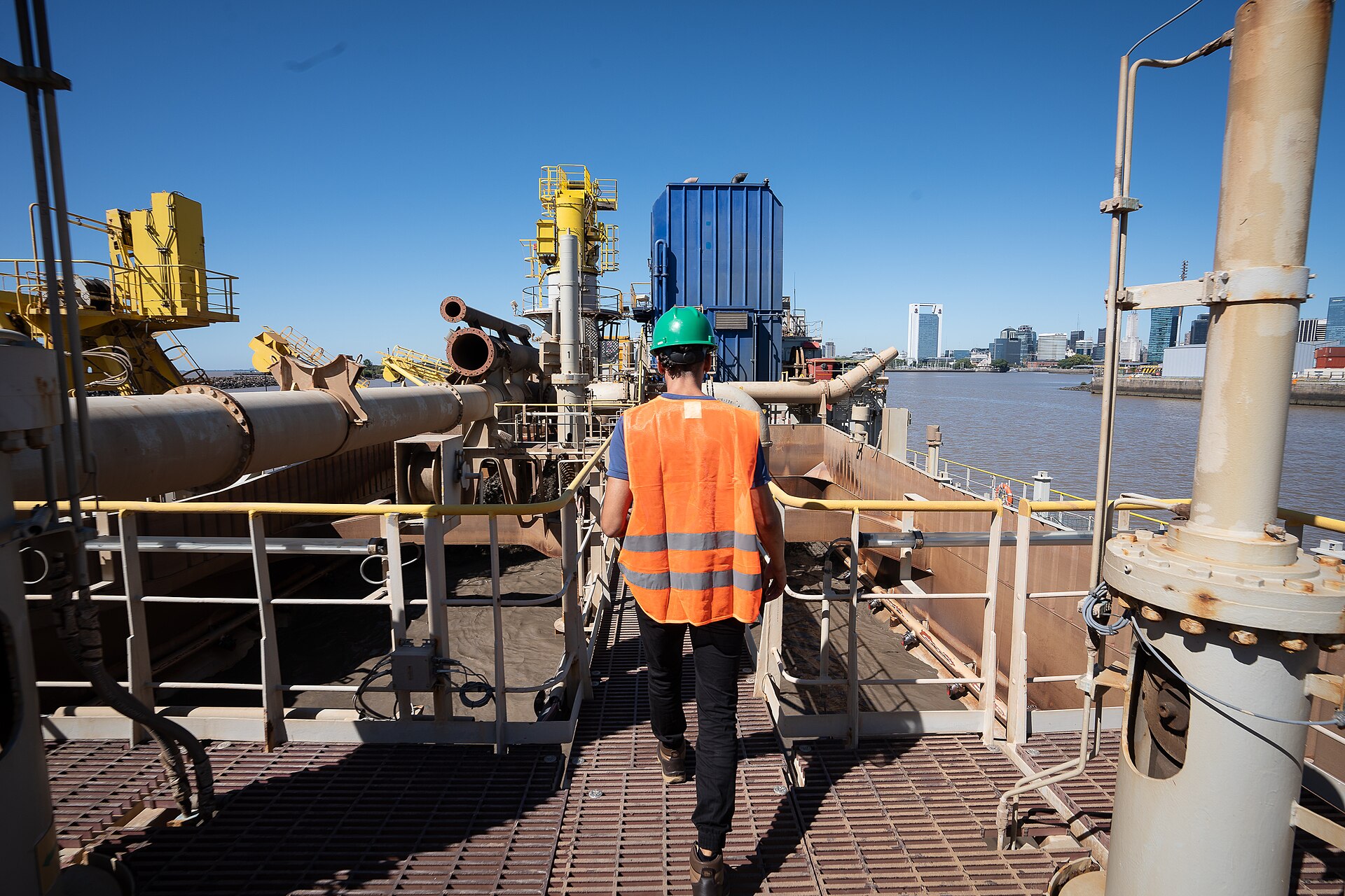 A worker in an orange vest and green helmet walks along the deck of a dredger at the port of Buenos Aires, with the river and city skyline beyond
