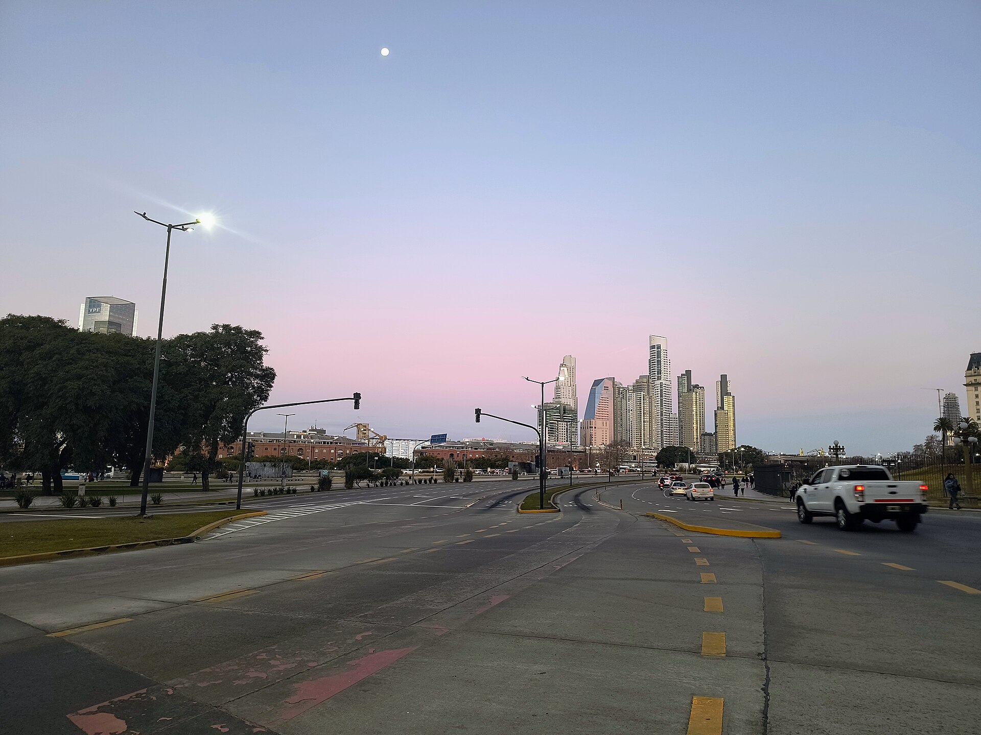 Glass towers of the Puerto Madero district rise over a wide avenue in Buenos Aires at dusk with a pink sky and a white pickup truck