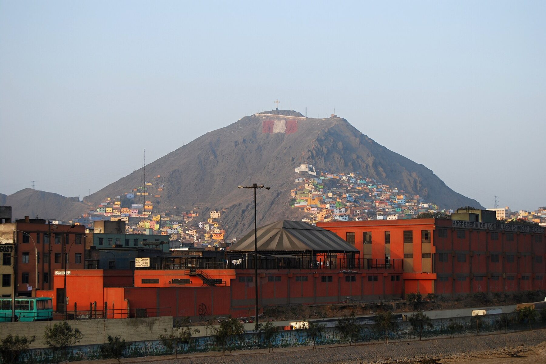 Brightly painted houses covering the lower slopes of Cerro San Cristóbal, a bare hill topped by a cross, behind orange buildings in Lima