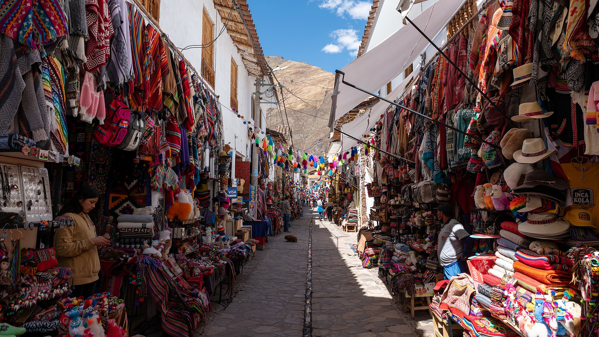A narrow stone street in Pisac lined with stalls selling colourful woven textiles, hats and bags, with a mountain in the background