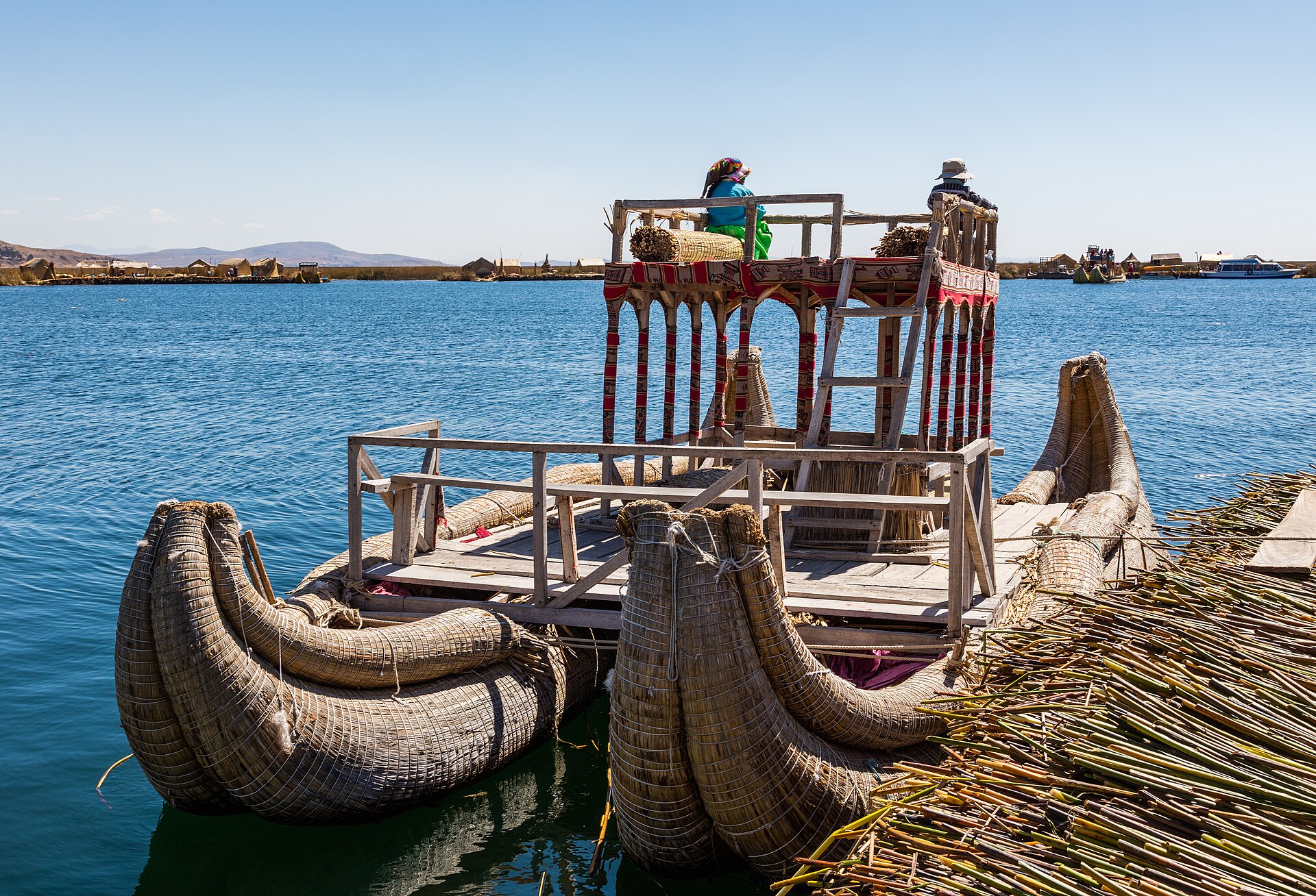 A large reed boat with a wooden upper deck moored at a floating reed island on the blue water of Lake Titicaca, with two people seated on top