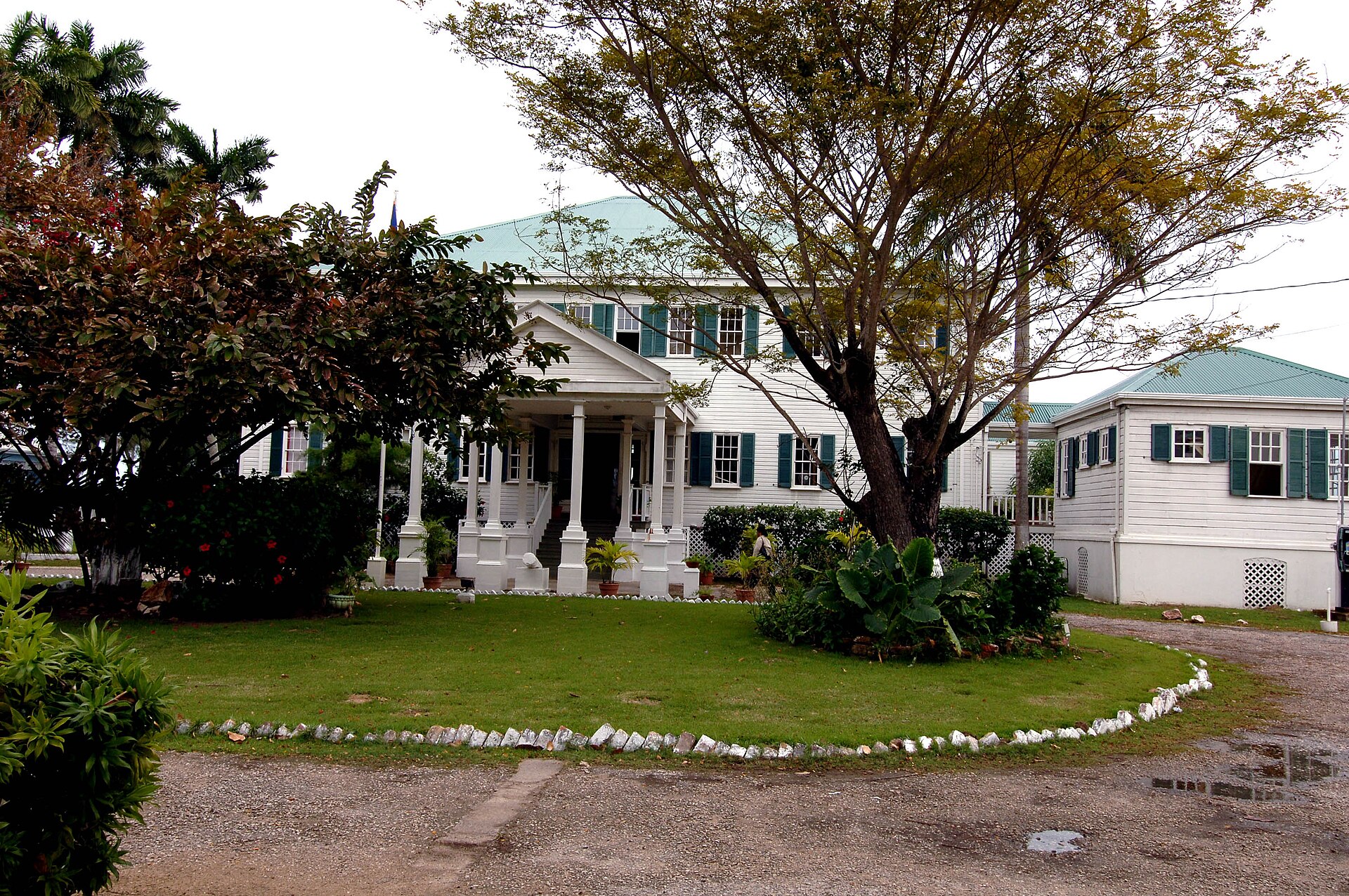 White colonial wooden house with a pillared porch and green roof behind a lawn, in Belize City