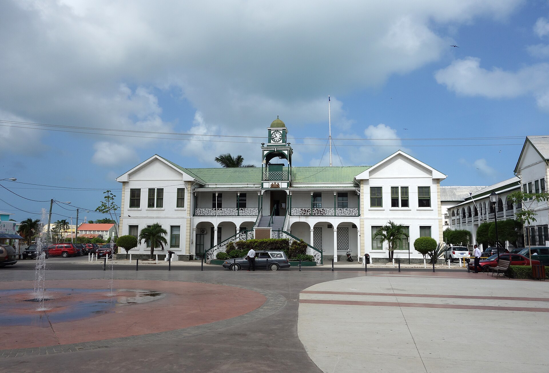 White two storey courthouse with a green roof and clock tower on a plaza with a fountain