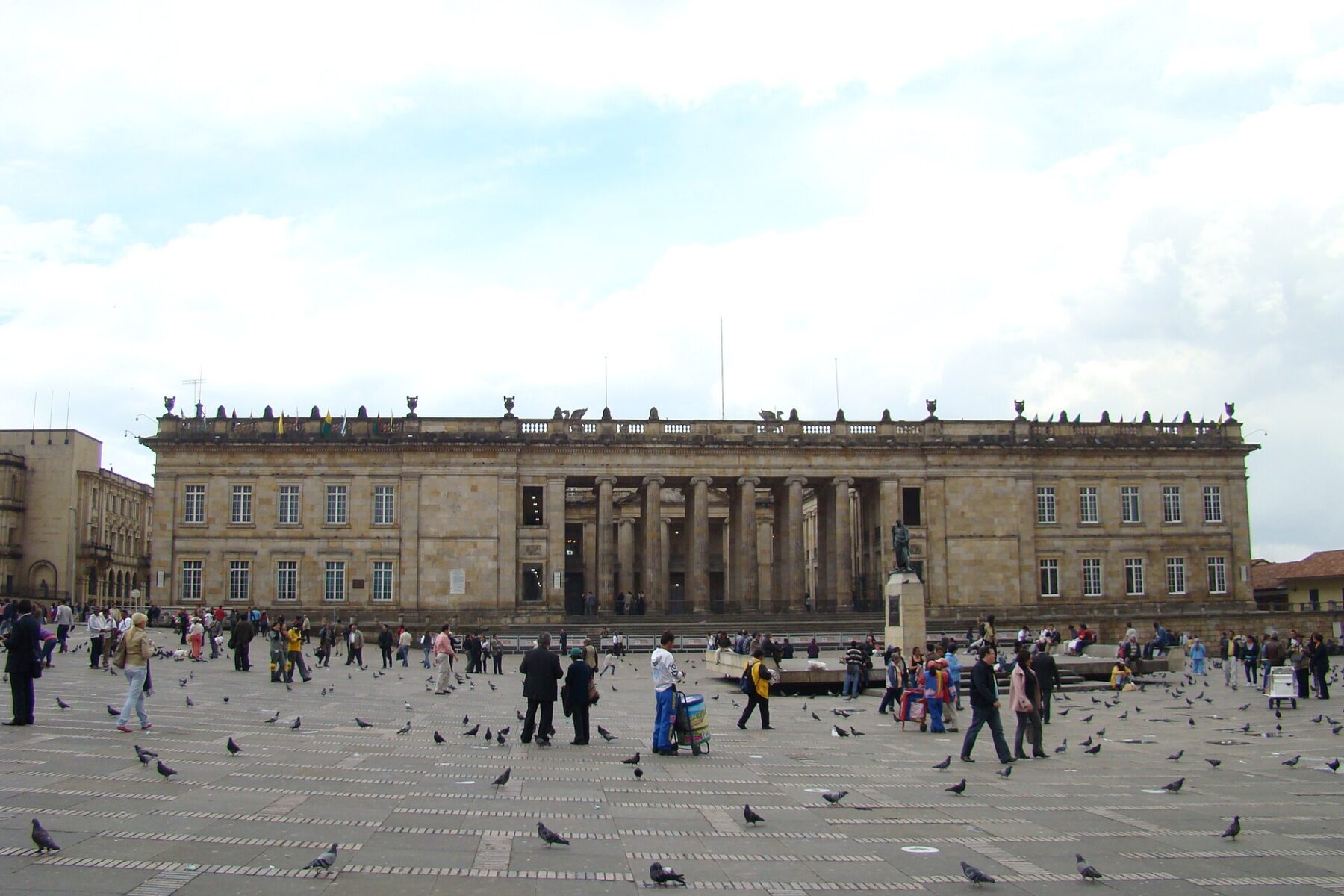 The columned stone facade of the Capitolio Nacional on Bolívar Square in Bogotá under a cloudy sky, with pedestrians and pigeons in the square