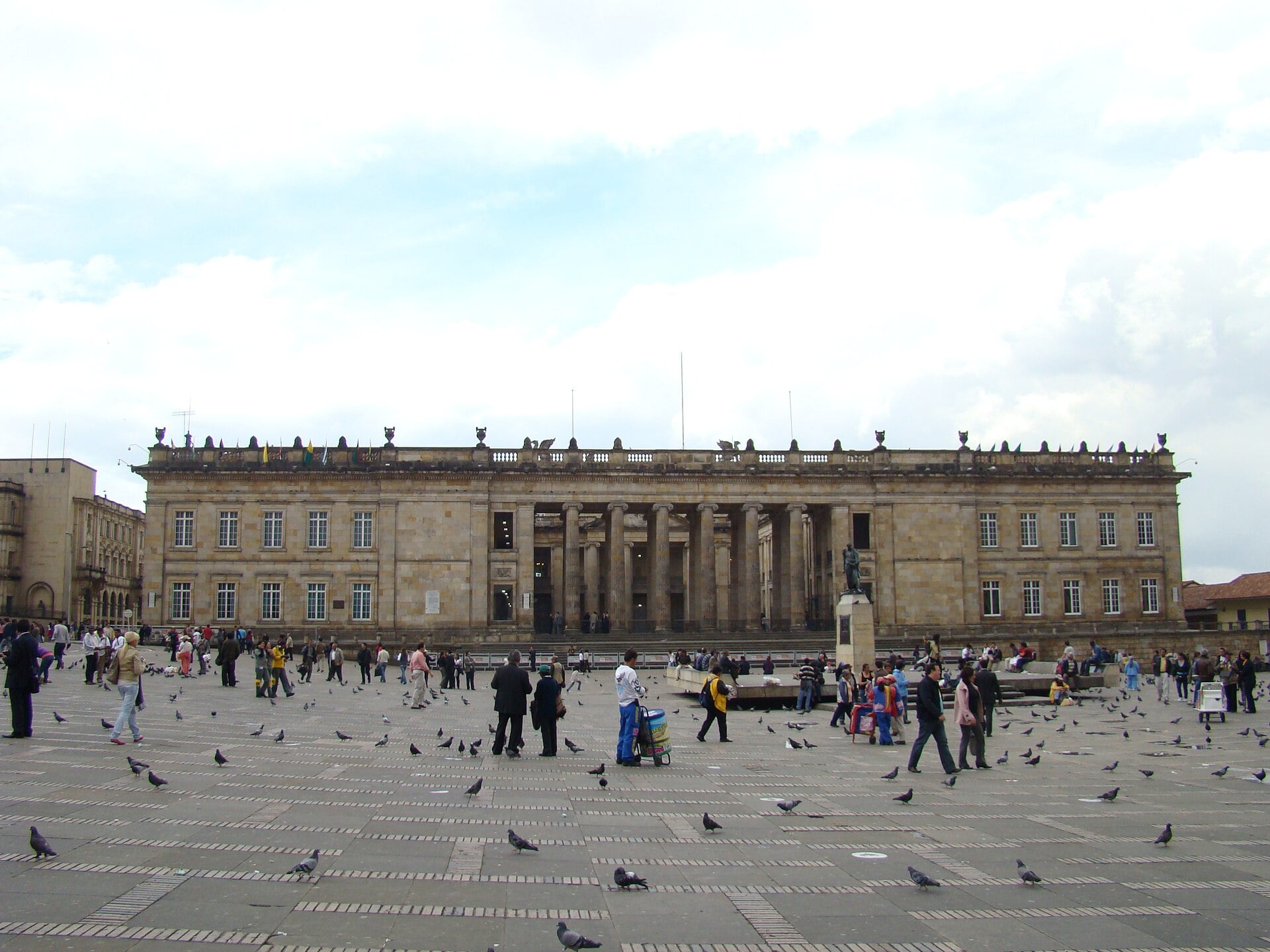 The columned stone facade of the Capitolio Nacional on Bolívar Square in Bogotá under a cloudy sky, with pedestrians and pigeons in the square